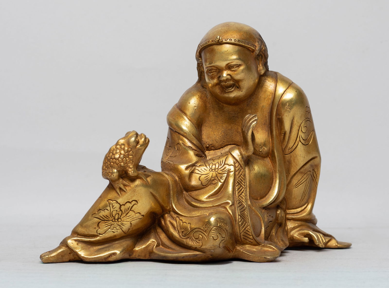 Chinese Gilt Bronze Sculpture of Luohan: Cast as Liuhai seated looking at a toad on his right knee, wearing a loose fitting robe incised with flowers. Approx. 7*4.9*3.3 inch. The Estate of the Store Closed Sale, Los Angeles. 2022.