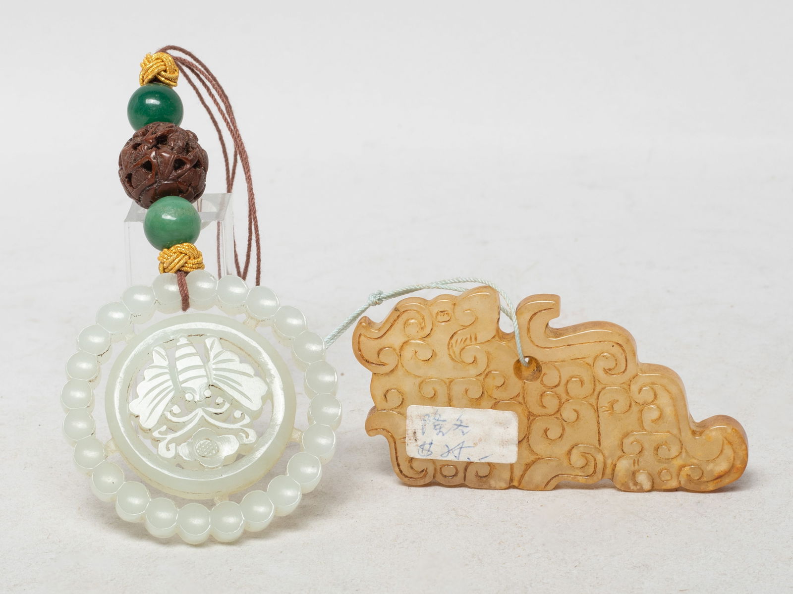 Two Chinese Jade Carved Pendants: The russet jade pendant carved of archaistic beast form, the white jade pendant of circular shape, reticulated with a butterfly and a flower with beaded frame, with a nut bead and two stone beads. App