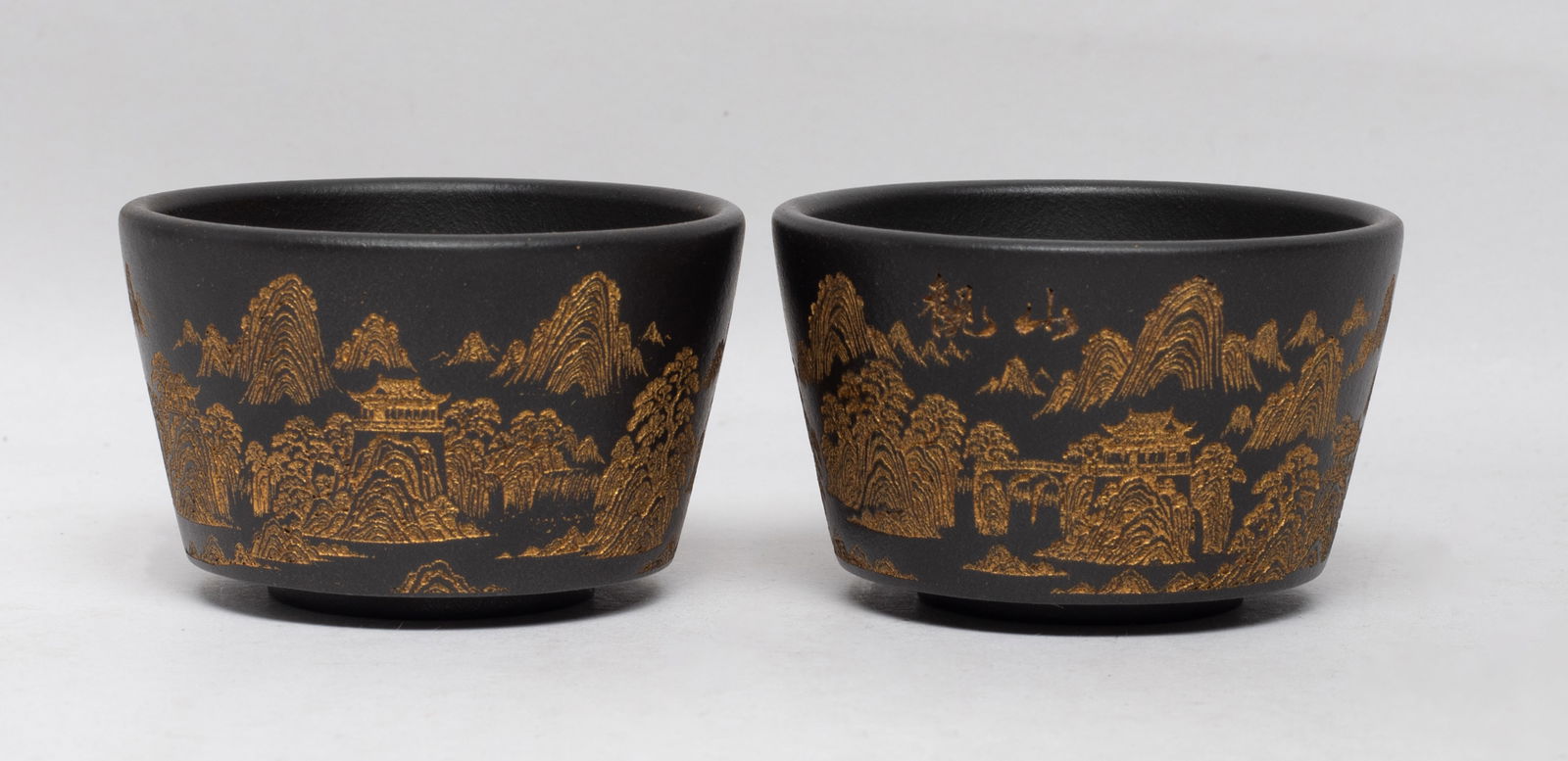 Two Chinese Zisha Clay Tea Cups: Each cup exterior incised and gilded with continuous mountain landscape scene, the bottom with a circular mark. Approx. 2.6*1.6 inch (each).