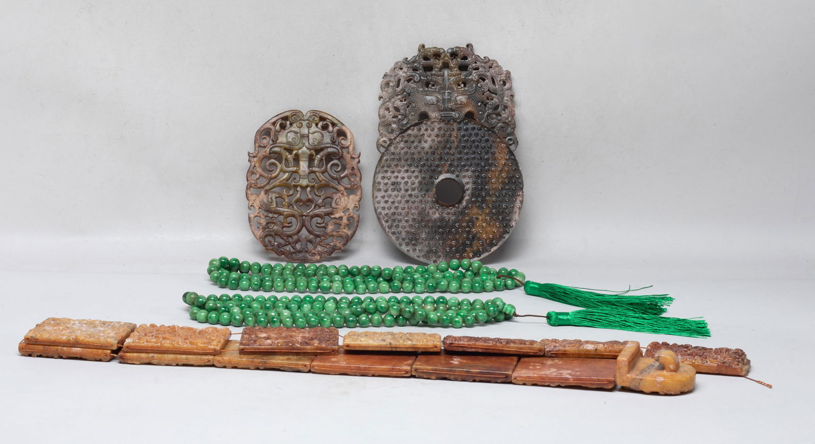 Chinese Export Stone Carvings, Colored Necklaces: Including two colored long bead necklaces, two stone carvings carved with archaistic beasts, masks or a scene of figures battling with beasts, and a stone belt carved with chi dragons among foliage.