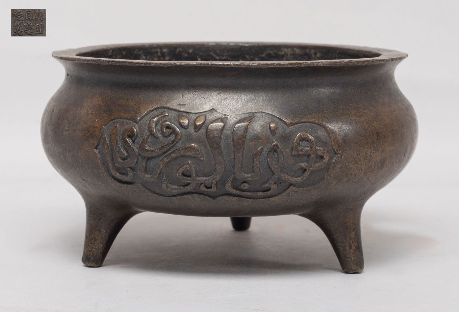 Collectible Chinese Bronze Cabinet Censer: Of compressed form rising from three tapered feet to a waisted neck and a flared rim, the exterior cast with two ruyi head shaped medallions of stylized characters, the bottom inscribed with a six-cha