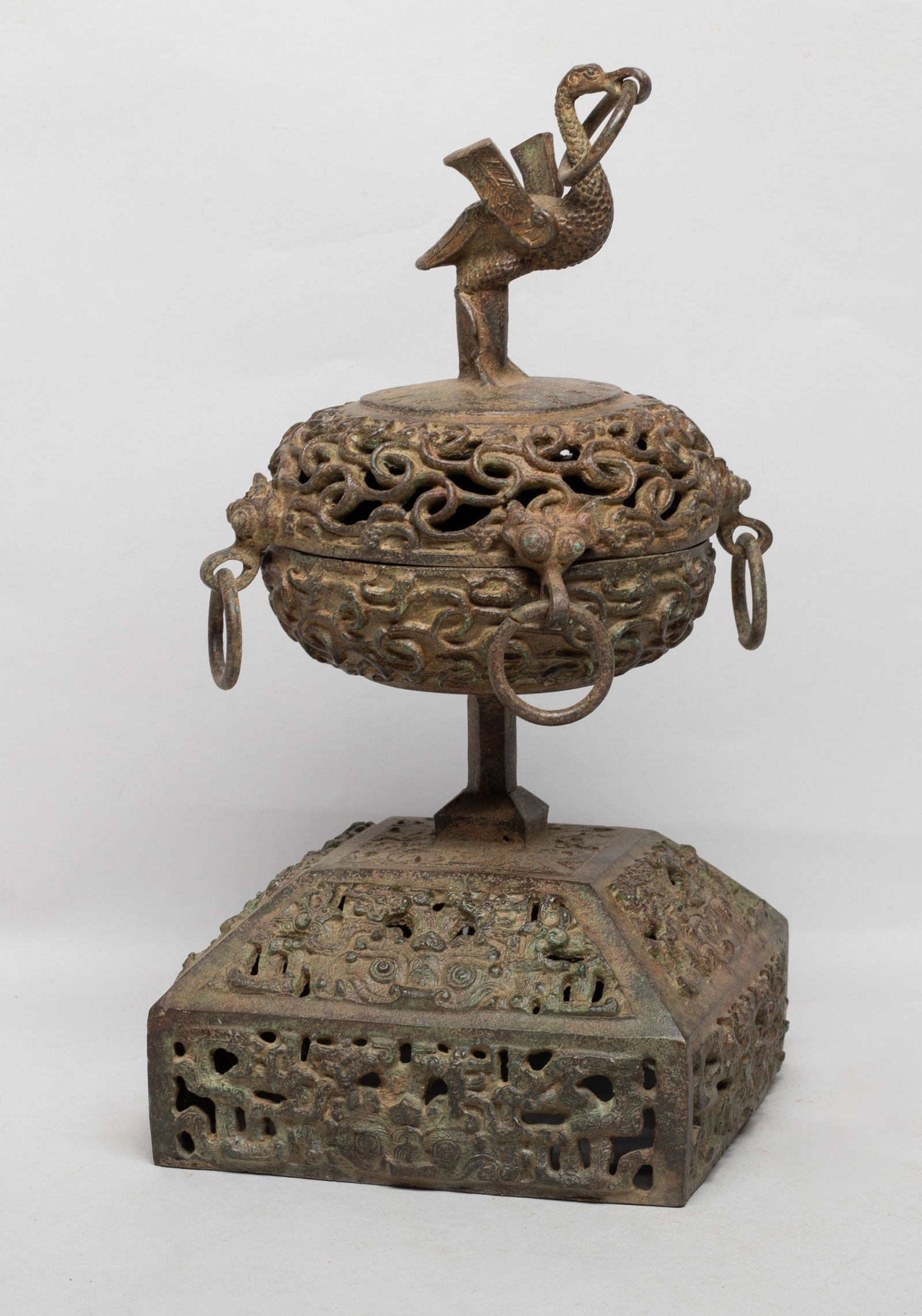 Chinese Bronze Covered Censer: The compressed globular body cast with intertwined archaic dragons, flanked by four mask handles suspended with loops, surmounted with a bird finial, all supported on a square base reticulated with ta