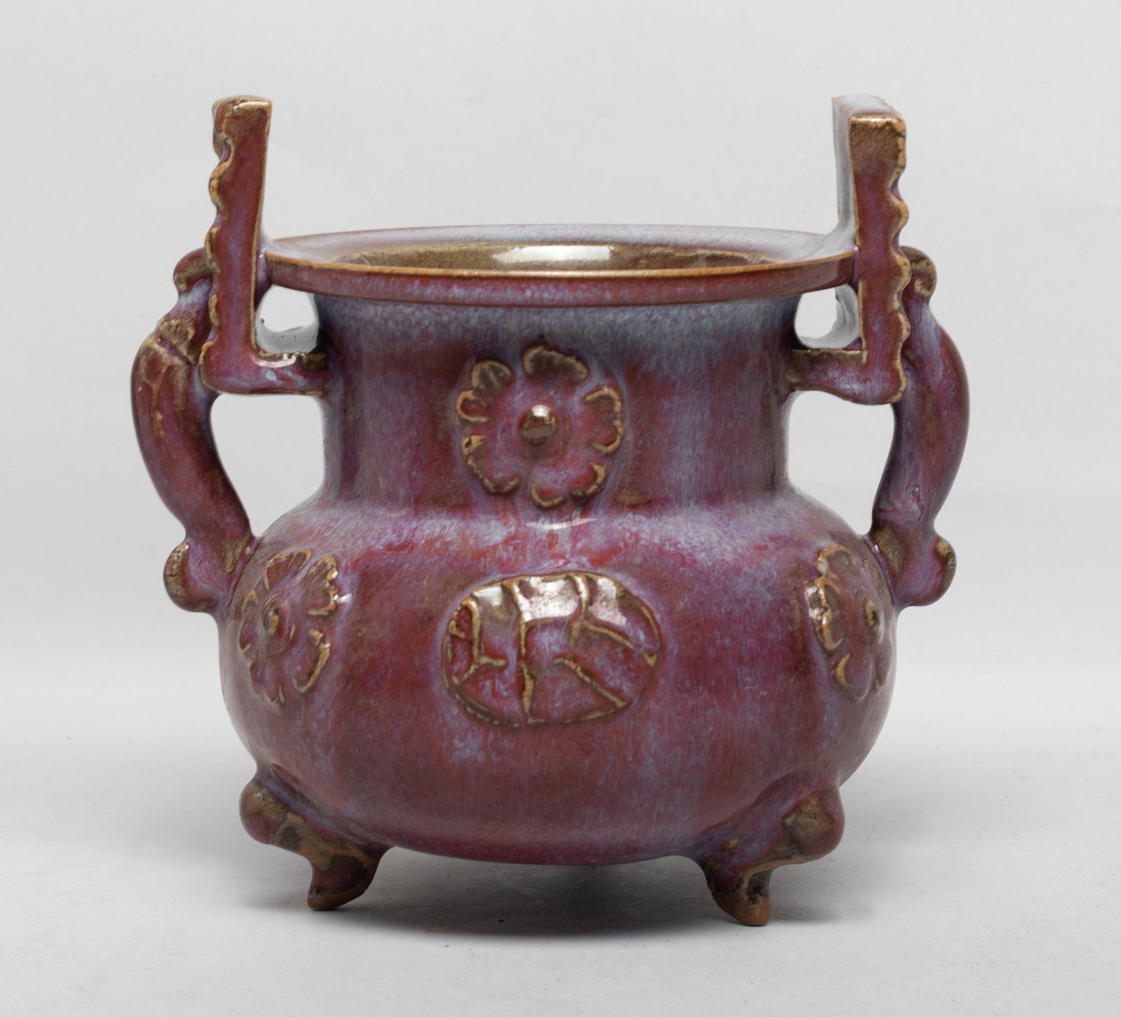 Chinese Jun Ware Porcelain Cabinet Censer: The compressed globular body rising from three short legs to a cylindrical neck and a flat rim, flanked with two handles, the exterior carved with stylized flowers, the exterior covered in purple red