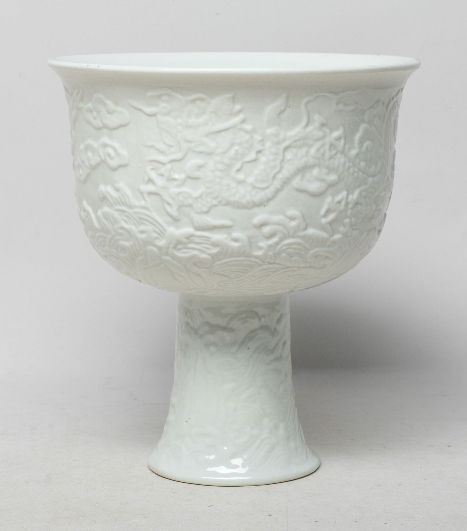 Large Chinese Molded Porcelain Tall Cup: The cup exterior carved with dragons sinuous amongst scrolling clouds above foaming waves and rocks, overall covered in even white glaze, the tall foot inside incised with a seal mark. Approx. 6.7*6.1