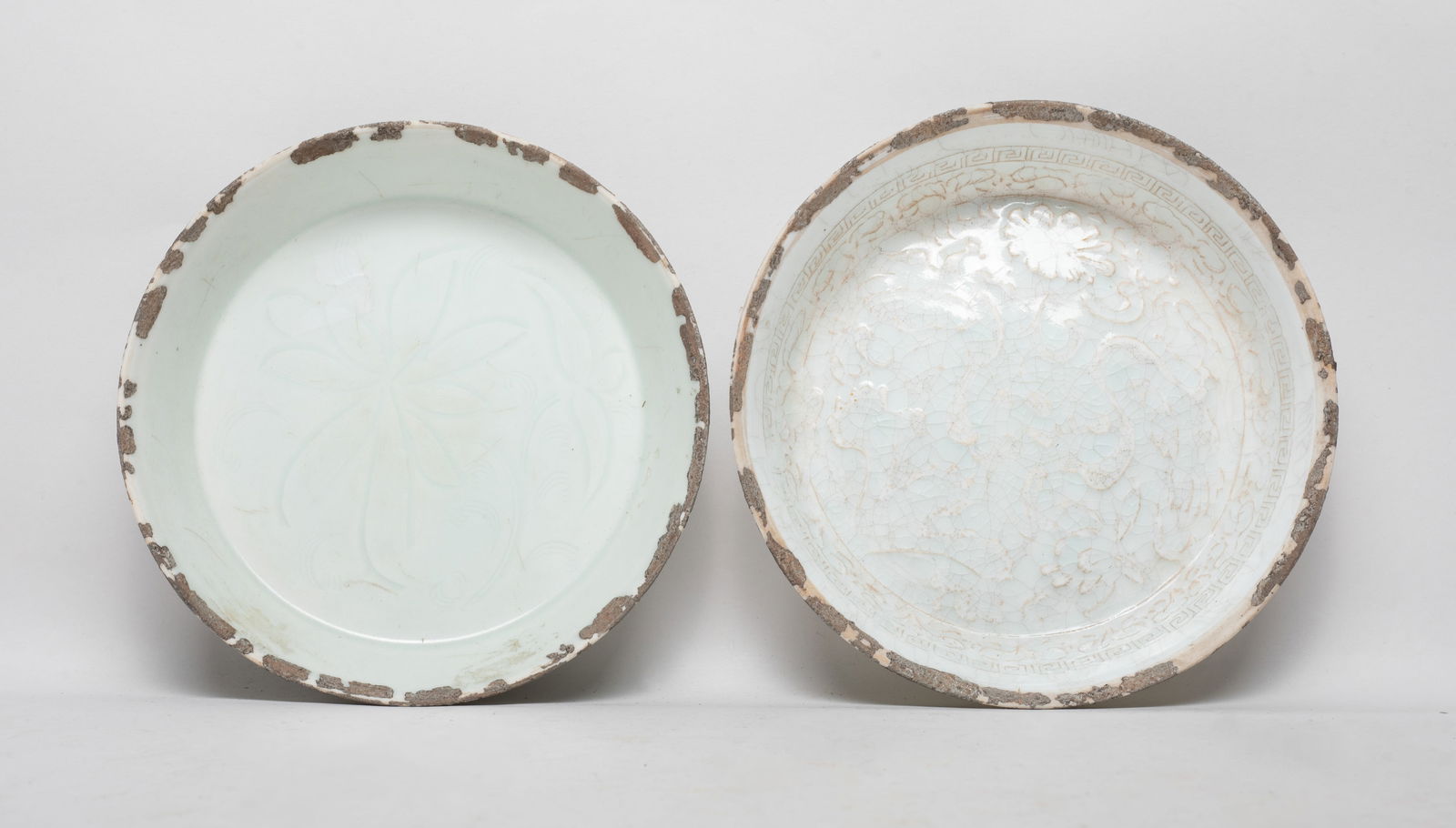 Two Chinese Hutian Type Porcelain Deep Plates: One plate interior carved with dense foliage framed by a fret border, overall covered with crackled bluish white glaze, the other plate interior incised with large blooms, overall covered with soft ce