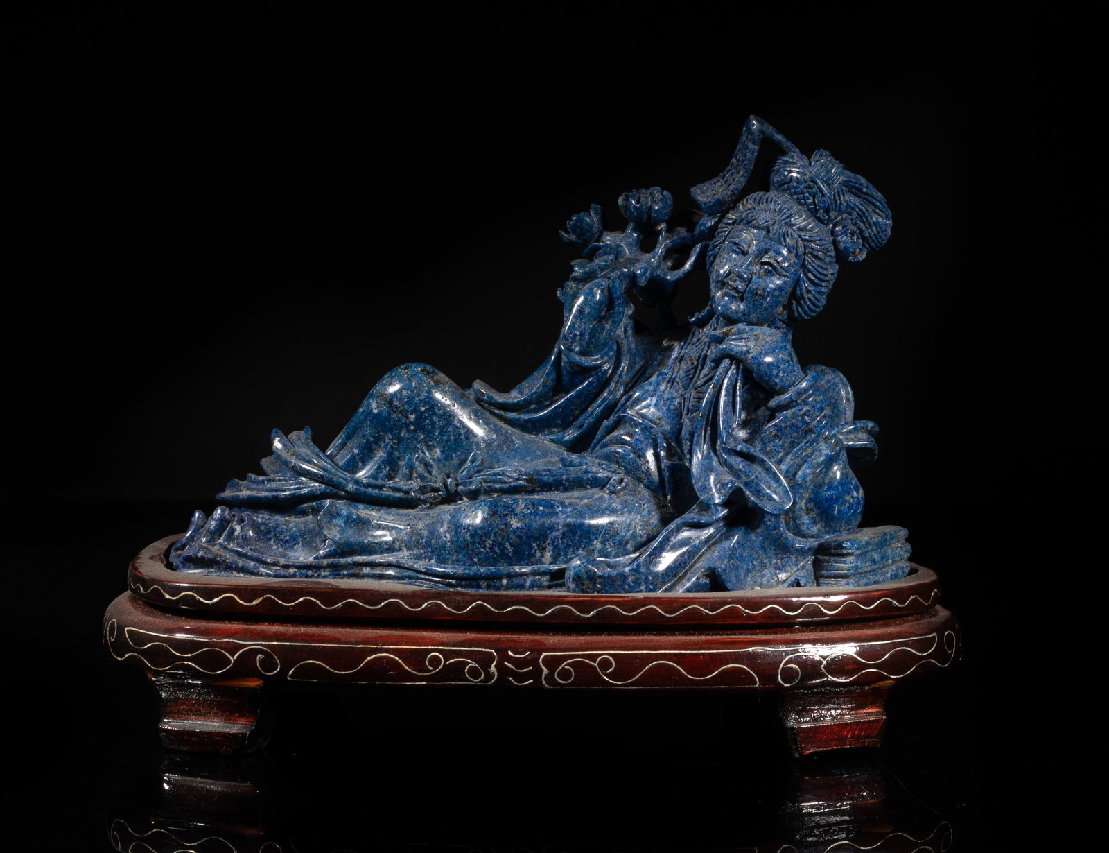 Large Chinese Rich Blue Lapis Sculpture: Carved as an elegant woman lying on her left side, the left hand grasping a handkerchief and right hand holding flowers, with a wood stand. Approx. 8*3.6*5.7 inch (w/ stand). The Estate of the Los