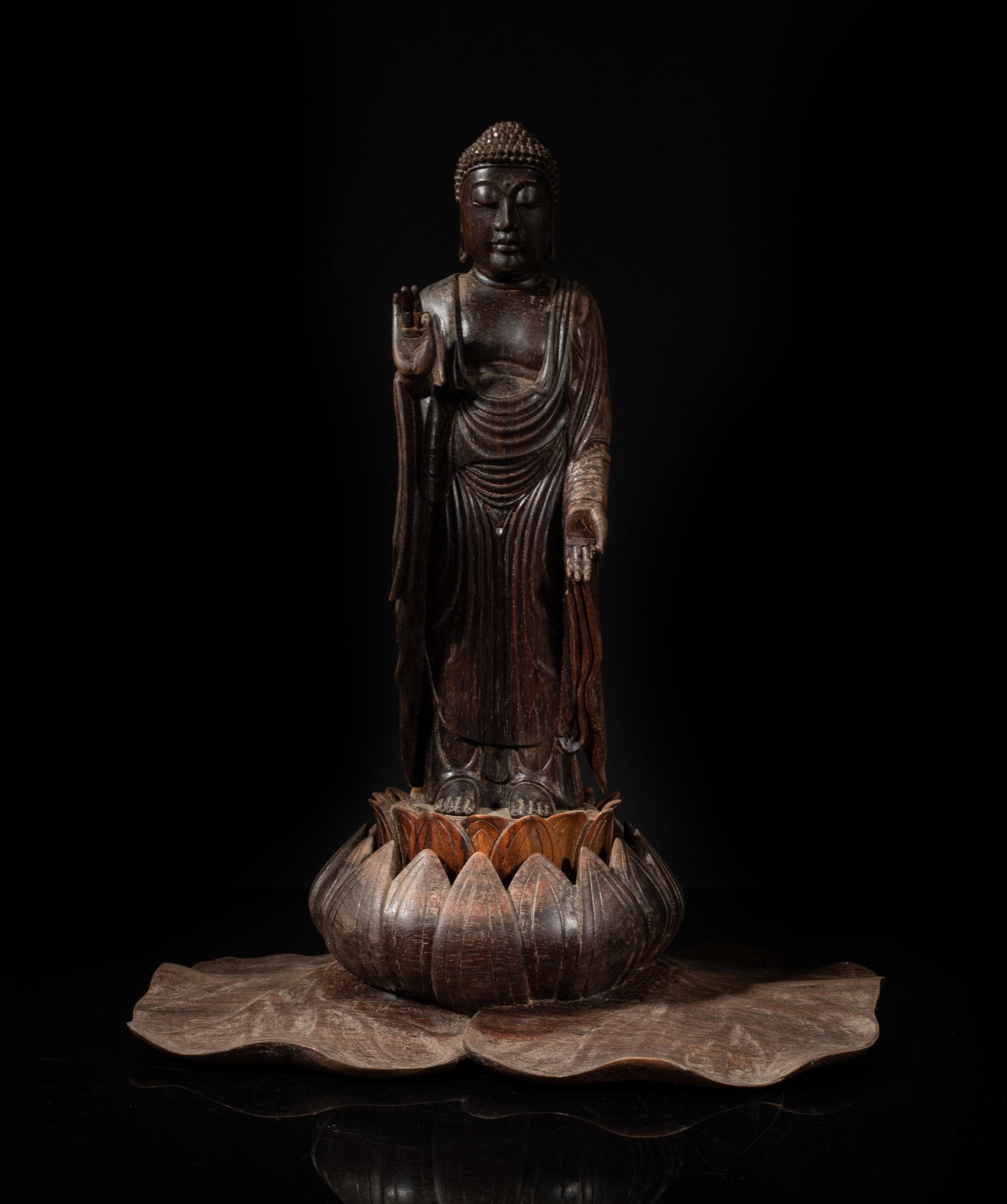 Tall 19th South Asian Rose Wood Buddha on Lotus: The Buddha carved standing on a lotus with a large lotus leaf base, dressed in long loose-fitting robe, the hands in vitarka mudra pointing to opposite directions, the rounded face with serene