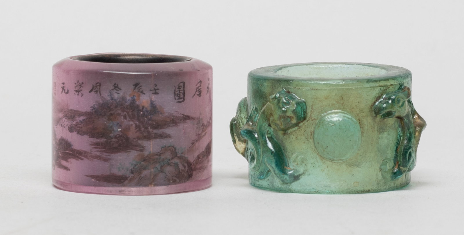 Two Rare Chinese Glass Thumb Rings: One pink glass thumb ring decorated with continuous landscape scene, the other blue-green glass thumb ring carved in high relief with a pair of chilong pursuing a ball. Approx. 1.6*1 inch (larger).