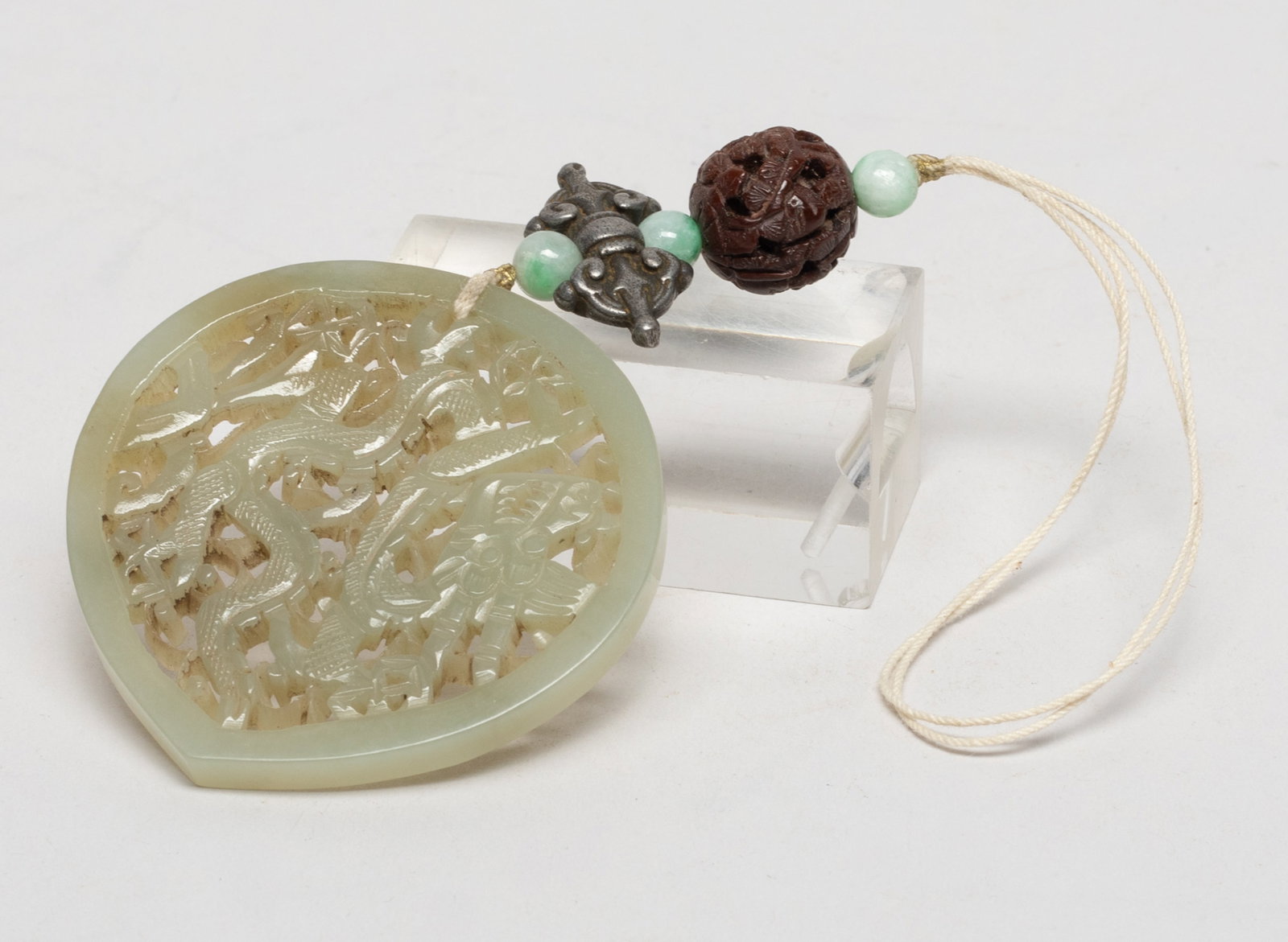 Estate Chinese Export Jade Pendant: Reticulated with a dragon sinuous amidst foliage, with three stone beads, a nut bead carved with figures, and a metal toggle. Approx. 2.2*2.2 inch (jade size). The Estate of Downtown, Los Angeles.