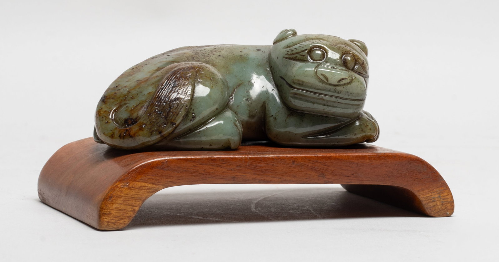 Chinese Russet Jade Beast w/ Wood Base: The imposing animal carved recumbent with wide nostril and large bulbous eyes beneath bushy brows, the accentuated spine terminating with the large bushy tail sweeping across its right haunch, with