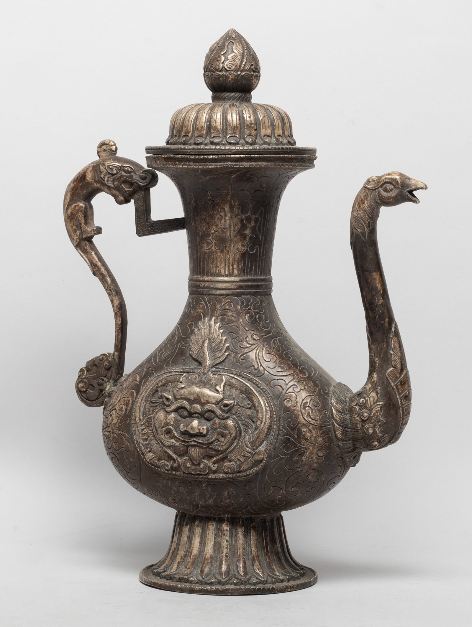 Tall Chinese Sino-Tibetan Silver Plated Pot: Of flattened pear shape rising from a splayed foot to a flared neck, either side carved in raised relief with a stylized mask against foliage ground, with beast form handle and spout, the lid with a