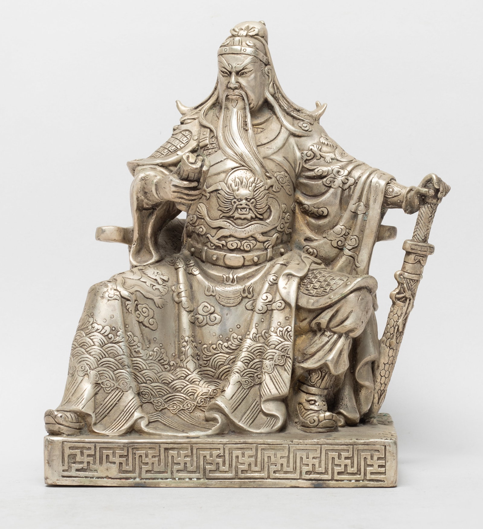 Vintage Chinese Silver Tone Sculpture of Kuanti: Cast seated on a beast mask chair, his left hand resting on a sword and right hand holding a book, wearing a long robe detailed with dragons pursuing a flaming pearl amidst clouds above waves.