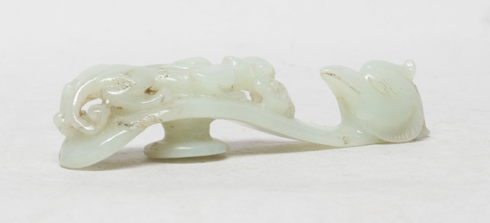 Estate Chinese jade Carved Belt Buckle: Of curving thin ovoid section terminating in a hook rendered as a phoenix head peering at the undercut and reticulated chilong clasping a sprig of lingzhi, the underside with a circular boss. Approx.