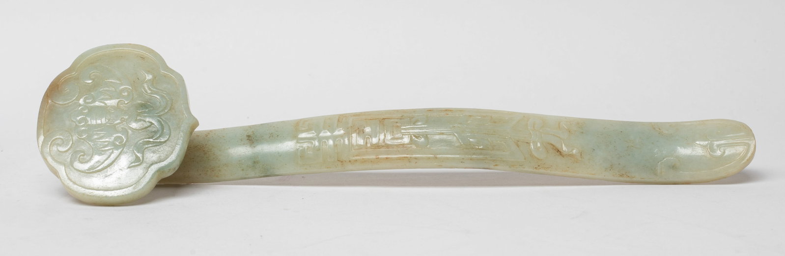 Chinese Jade Carved Sculpture of Ruyi: The ruyi head carved with a bat grasping a beribboned ball in mouth, the arched shaft carved with archaic motif, the jade of pale celadon tone with russet inclusions. Approx. 9*2.2*1.3 inch.