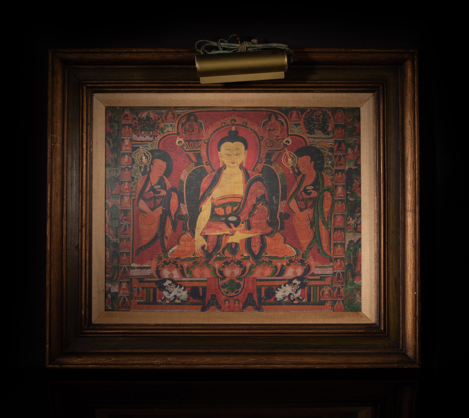 Chinese Export Wall Hanging Thangka: Depicts a buddha seated on a lotus base in a meditative posture, dressed in red and gold robes, flanked by two standing attendants, the frame with an integrated picture light. Approx. 28*24.5 inch (w/