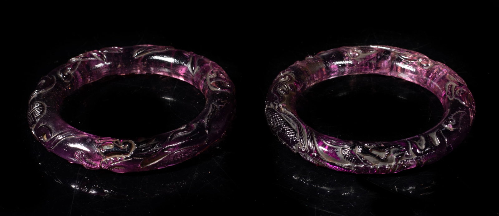 Two Chinese Amethyst Like Glass Bangles: Two Chinese amethyst-like glass bangles, carved with dragon, phoenix, figure and beast. Approx. 3.3 inch outer diameter, 2.3 inch inner diameter (each). The Estate of Kuo & Lee Art. HK. 1986.
