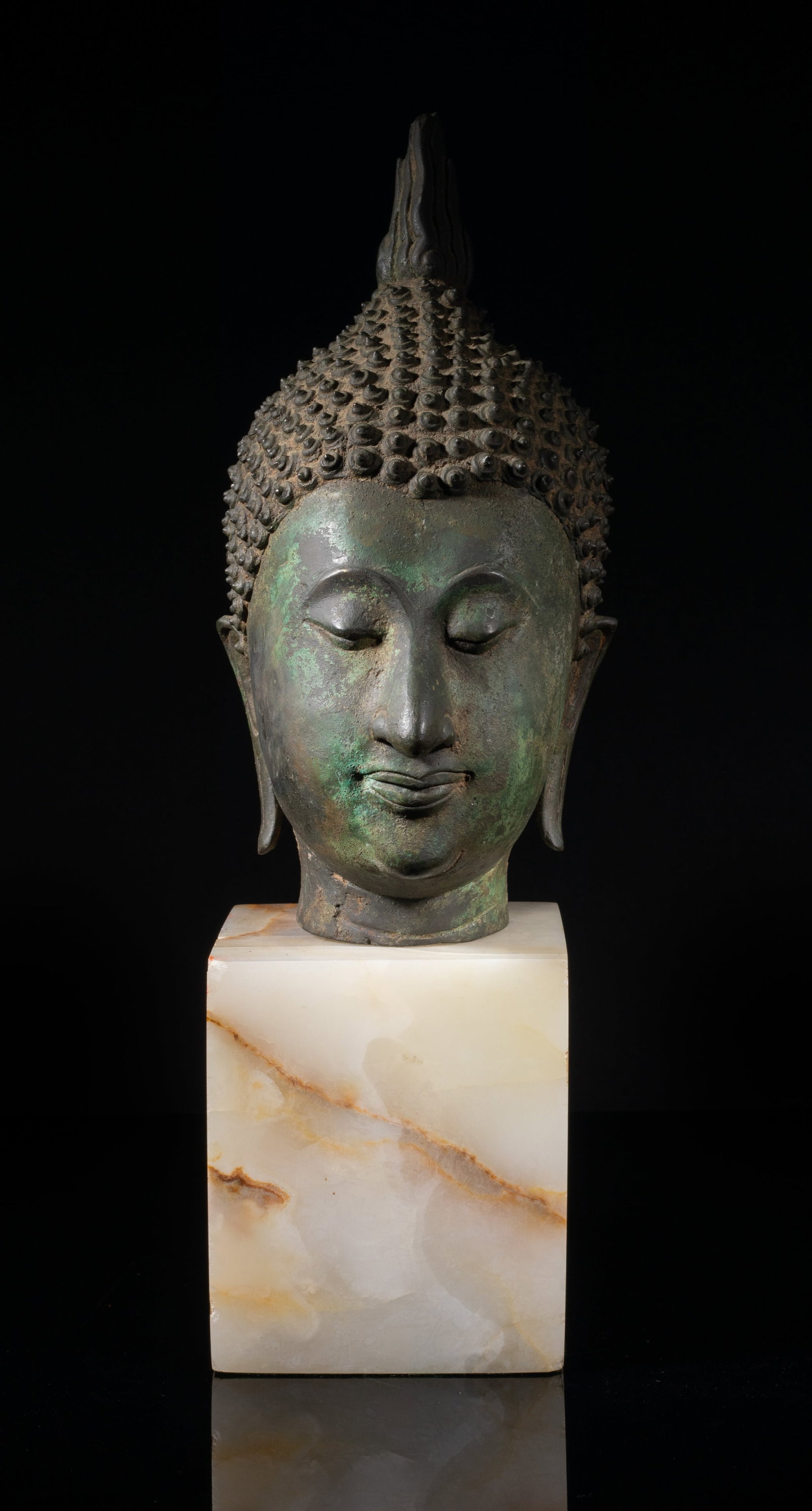 18th South Asian Large Bronze Buddha Head: The slender face modeled with downcast eyes and gentle smile, flanked by a pair of pendulous ears, with tight spiral curls covering the top of the head and a flame rising from the usnisa, with a stand