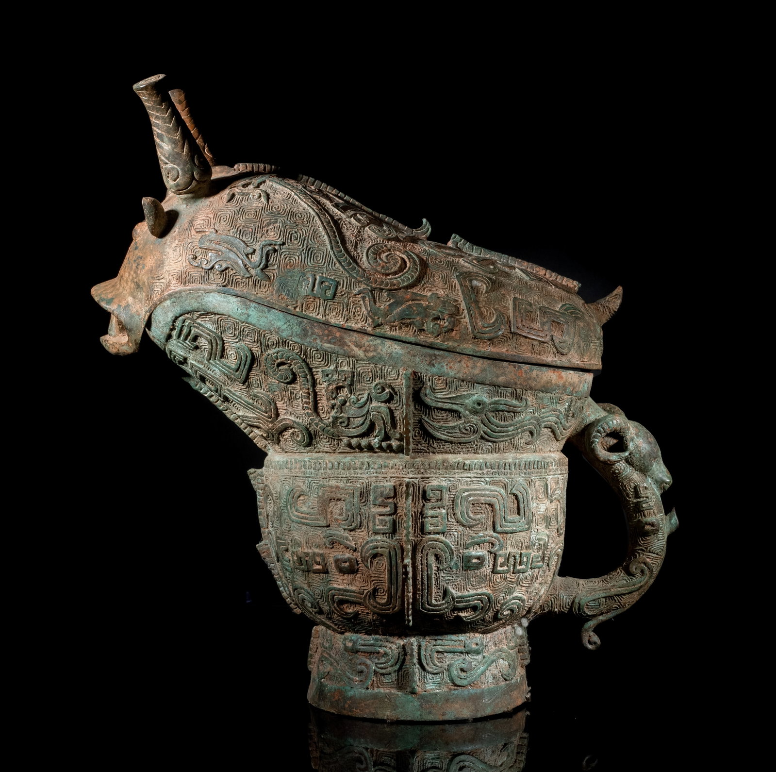 Massive Chinese Bronze Covered Vase: Of oval cross-section with a deep ascending spout, set on a cylindrical foot, the plain body cast with notched flanges and taotie masks, one loop handle issuing from a bovine head with rounded eyes an