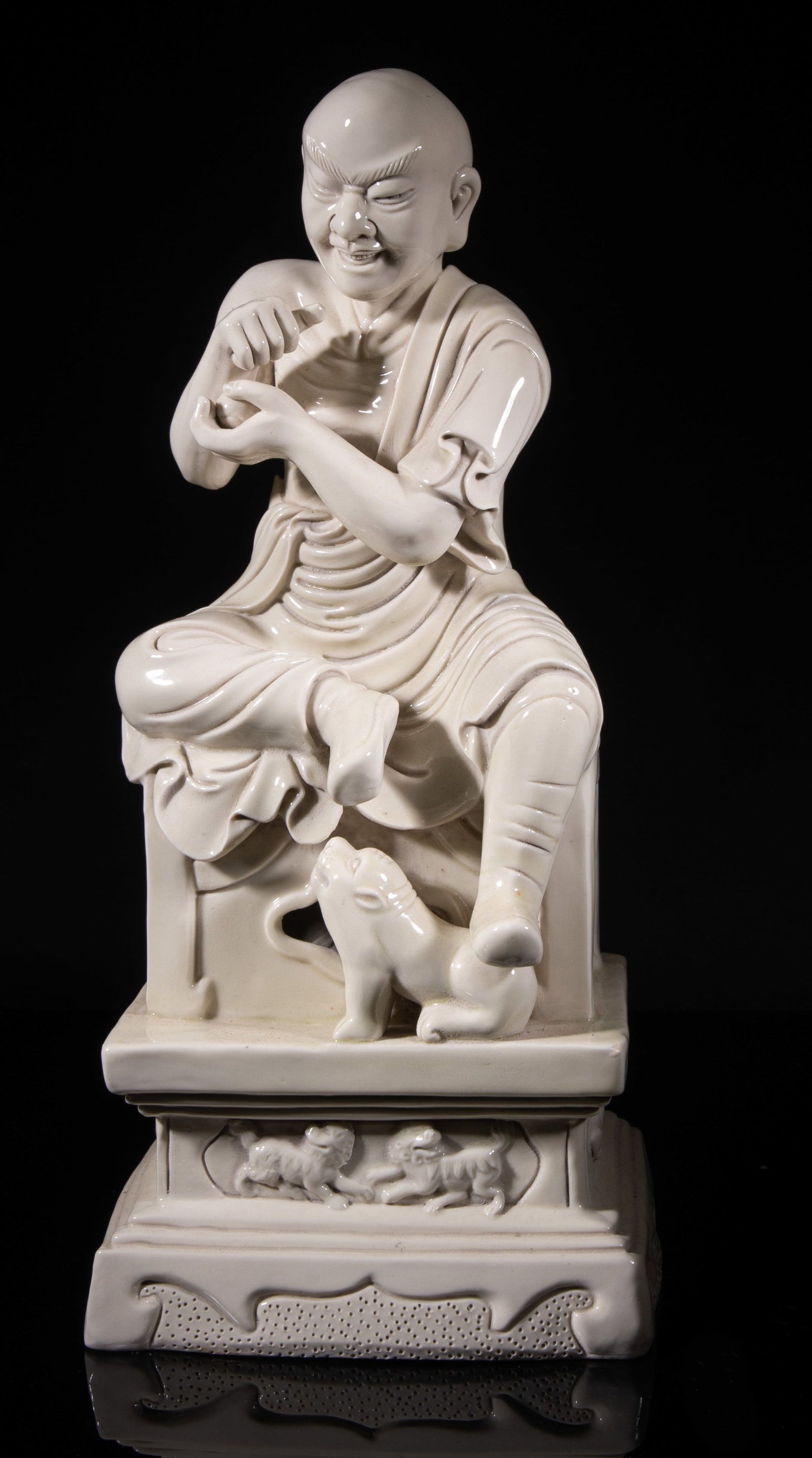 Chinese Blanc De Porcelain Sculpture of Luohan: The Luohan molded standing on a tall pedestal, carved with beasts and lotus, his left foot stepping on a crouching tiger, the hands holding a ball, the back incised with a gourd shaped mark and a seal