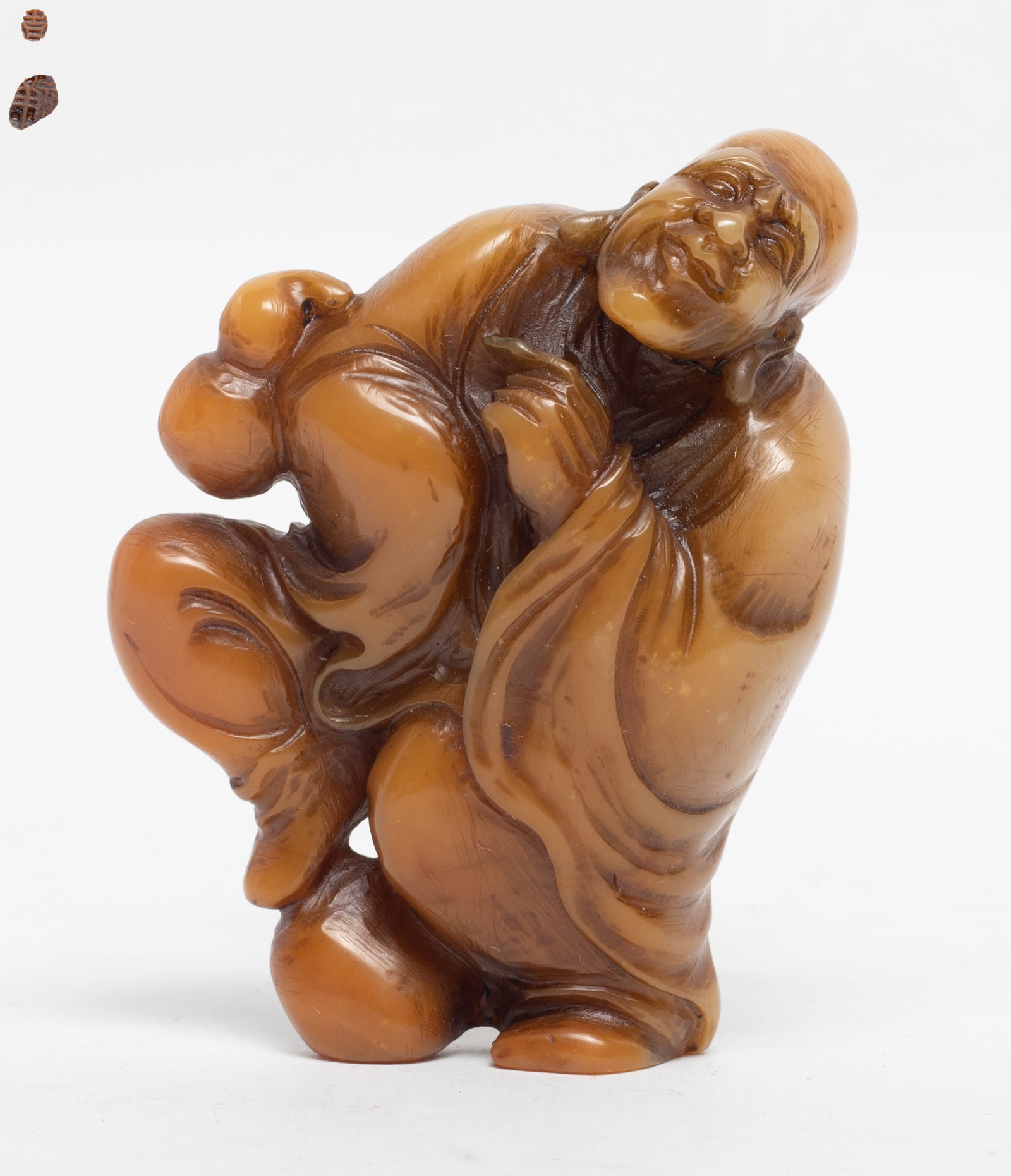 Chinese Shoushan Stone Carved Luohan: The figure in a seated position with a rounded back, depicts an elderly, bald monk with a serene meditative expression, the stone overall applied in an amber coloration, the base inscribed with a two-