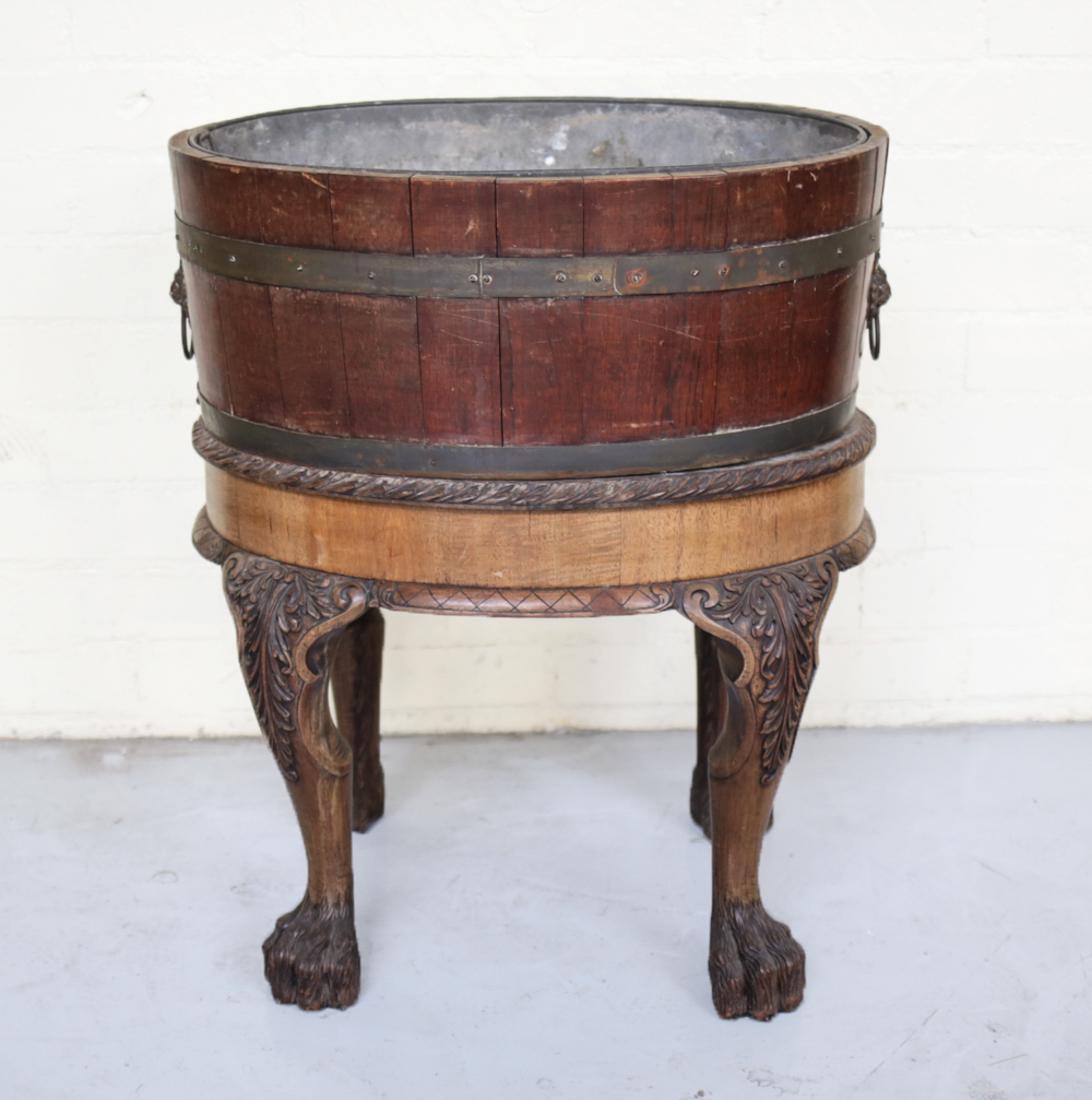 English Wine Cooler c1800: English Wine Cooler, with hand carved wood stand, tin insert. C1800. Oval barrel style container, bronze lion head handles. Stand has hand carved lion claw feet, foliate designs. The barrel is removab
