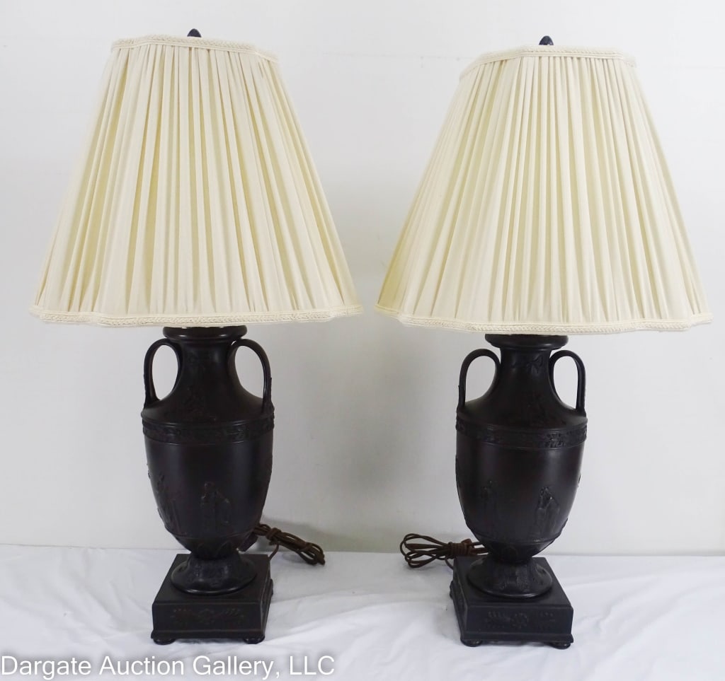 PAIR BLACK BASALT URN LAMPS: A pair of wedgwood style black basalt urn lamps with classic roman figures/scenes, each on four metal feet, and a double socket. These have been rewired. / Measures: 31.5" x 6" x 6" / Provenance: From