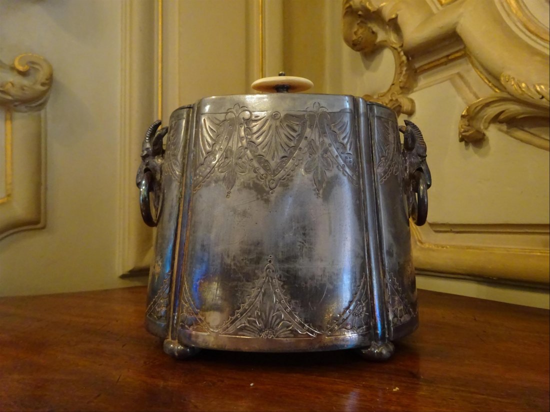 Argenterie: Large BOITE A THE en mtal argent, de section ovale, parties latrales flanques de ttes de bliers retenant des anneaux, frtel en ivoire.