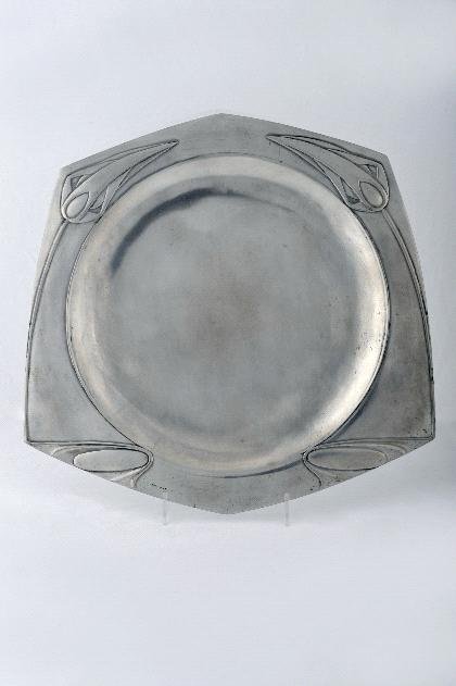Joseph Maria Olbrich, Zierteller, um 1902: Ausführung: Metallwarenfabrik Eduard Hueck, Lüdenscheid, Modell-Nr.1820. Zinn, poliert, Fahne mit reliefiert gegossenem, gegengleich angelegtem, streng stilisiertem Motiv sich neigender Blütenstän
