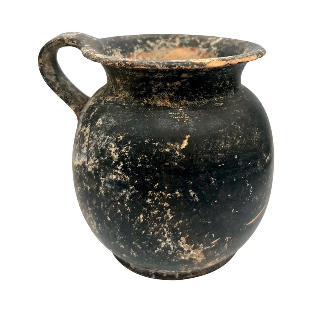 Greek black glazed oinochoe: Greek black glazed oinochoe, c. 5th - 4th Century BC, a drinking cup, the squat body with wide, flat bottom, flared mouth and wide looped handle. Size: 10.5 cm x 10 cm Some surface abrasions and depos