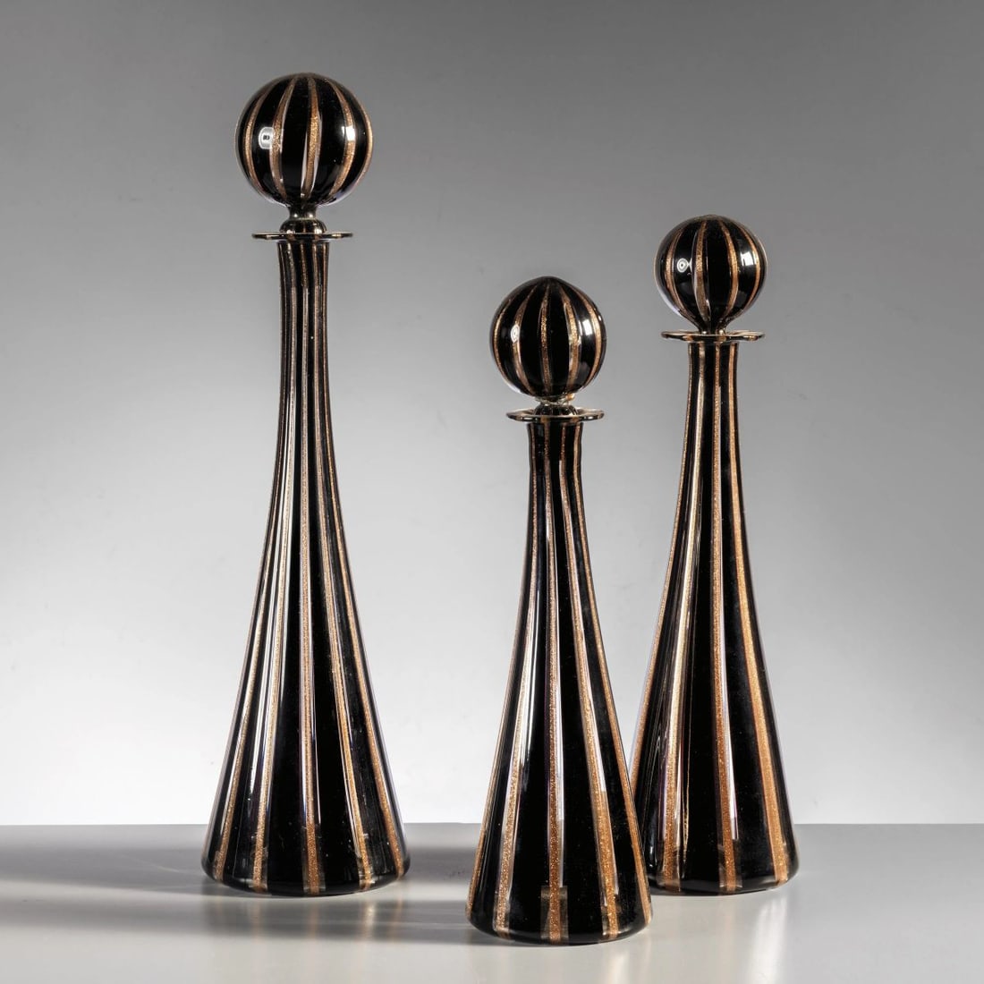 THREE BLACK AND GOLD GLASS BOTTLES: THREE BLACK AND GOLD GLASS BOTTLES TRE BOTTIGLIE in vetro nei toni del nero e dell’oro Produzione Italia, 1960 circa alt. cm 35 e diam. cm 10 alt. cm 29 e diam. cm 9 alt. cm 27 e diam. cm 8