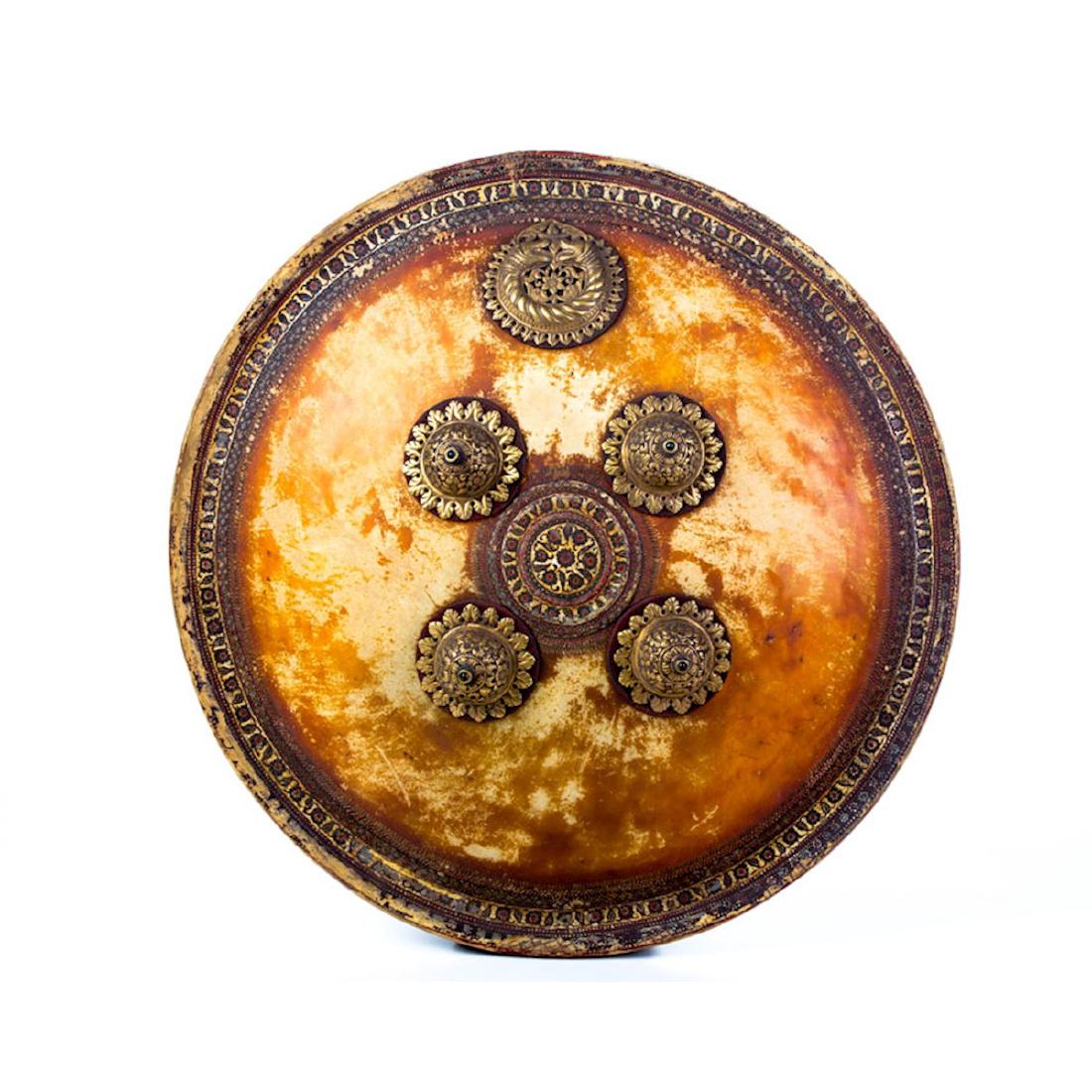 INDIAN HAND PAINTED DHAL SHIELD, 19TH C.: Light tan, translucent rhinoceros hide with delicately painted border and center. 4 ornate jeweled bosses and a 5th ornament depicting facing animal heads. The bosses are heavily gilded. diameter 17 7