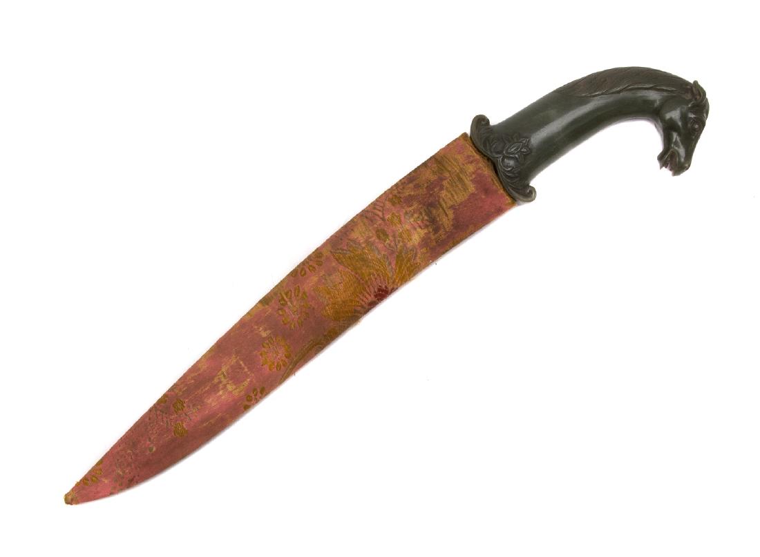 A MUGHAL STYLE HORSE-FORM JADE HILTED DAGGER: Recurved wootz steel blade with multiple fullers, the jade hilt with scroll quillons and carved stylised lotus leaf, the pommel in the form of a horse head; in its wooded scabbard covered with fabric.