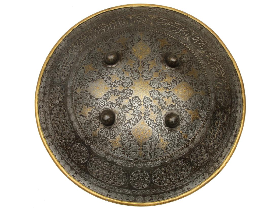 INDIAN INDO-PERSIAN ISLAMIC SHIELD: Large shield chiseled with floral motifs and Arabic inscriptions, decorated with gold koftgari and brass rim. Dimensions: Diameter: 18 3/8 in., (47 cm.)