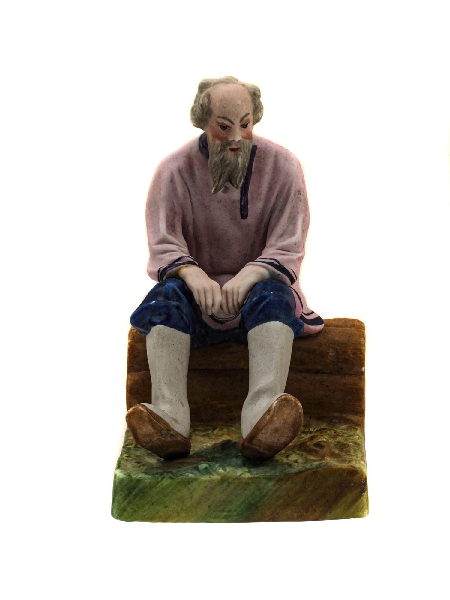 A RUSSIAN SOVIET DMITROV PORCELAIN FIGURINE 1920S: An early 20th-century porcelain figurine of the Dmitrov porcelain factory in Verbilki (formerly the Gardner factory) "A Seated Peasant". Depicts a gray-haired man in a pink shirt, blue pants, and high