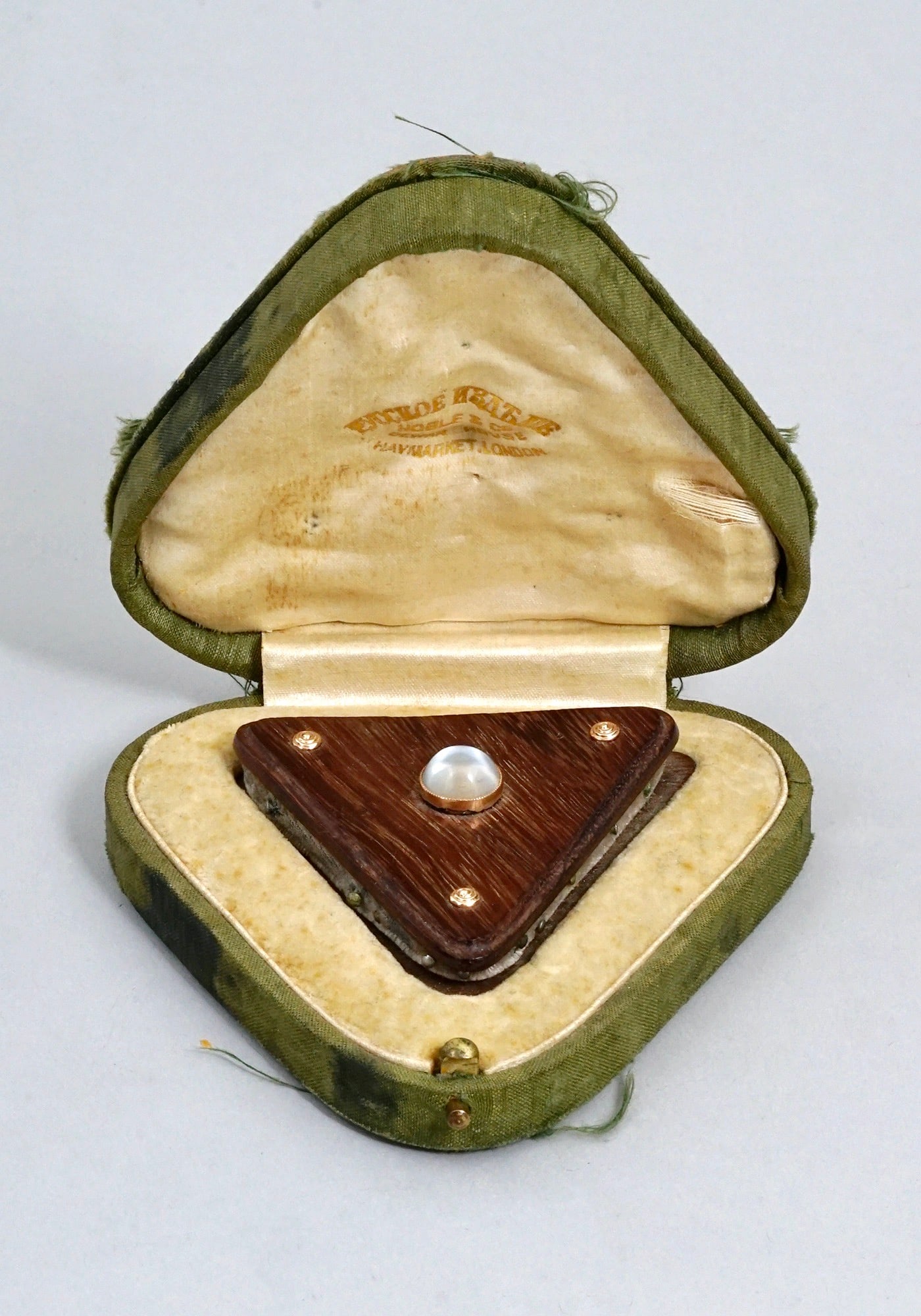 A CASED RUSSIAN WOOD NEEDLE HOLDER RETAILED BY NOBLE & CO., C. 1900: Russian needle case retailed by Noble & Co of London. Of a triangular form, made of wood with a velvet cushion between wooden parts. Set with a round cabochon-cut moonstone in a gilded frame. Comes in