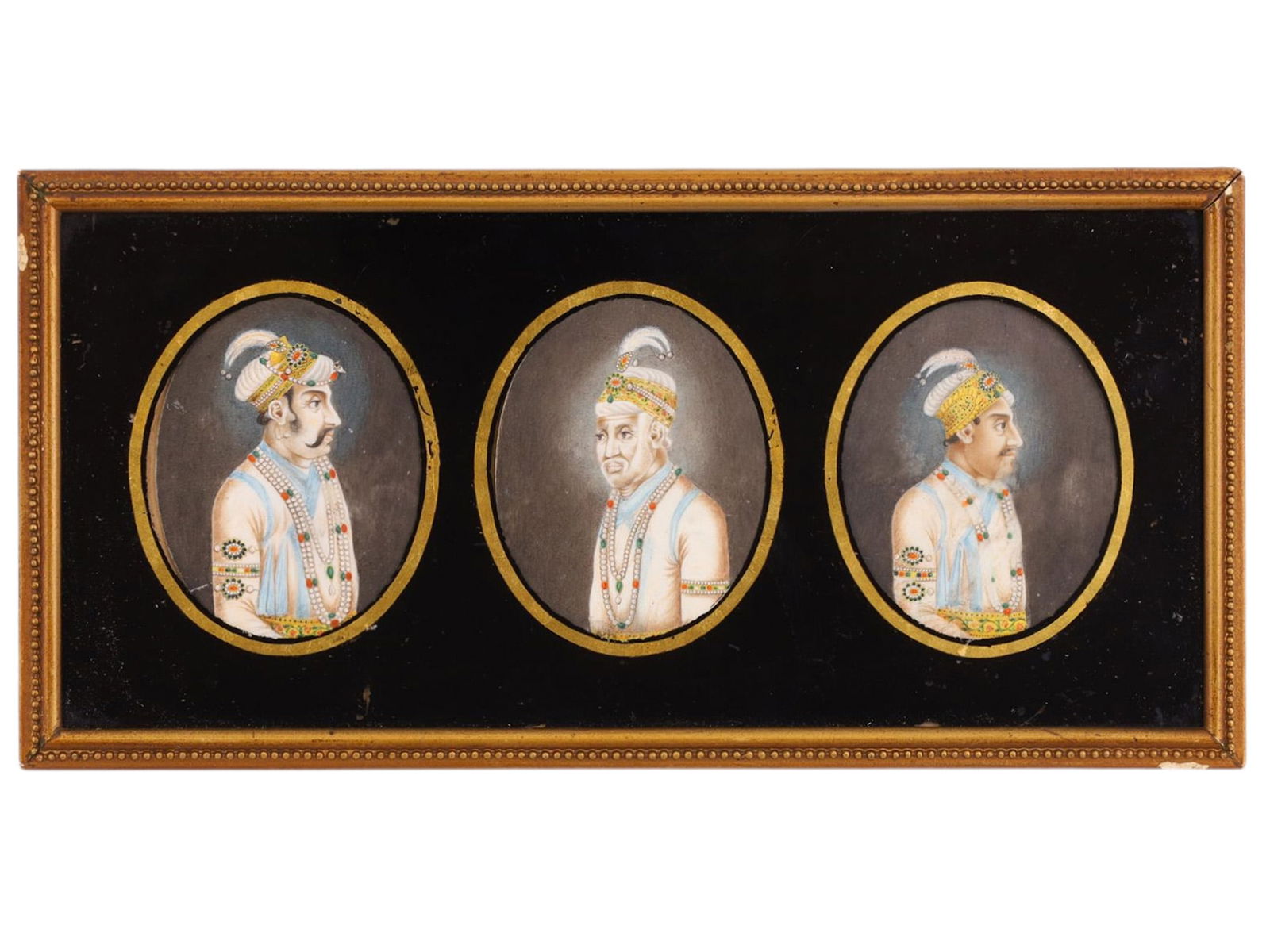 THREE INDO-PERSIAN MINIATURE PAINTINGS: Comprising three oval miniature portraits executed in opaque watercolor on paper, mounted within a single glazed rectangular frame against a black ground with gilt oval surrounds. Each miniature depic