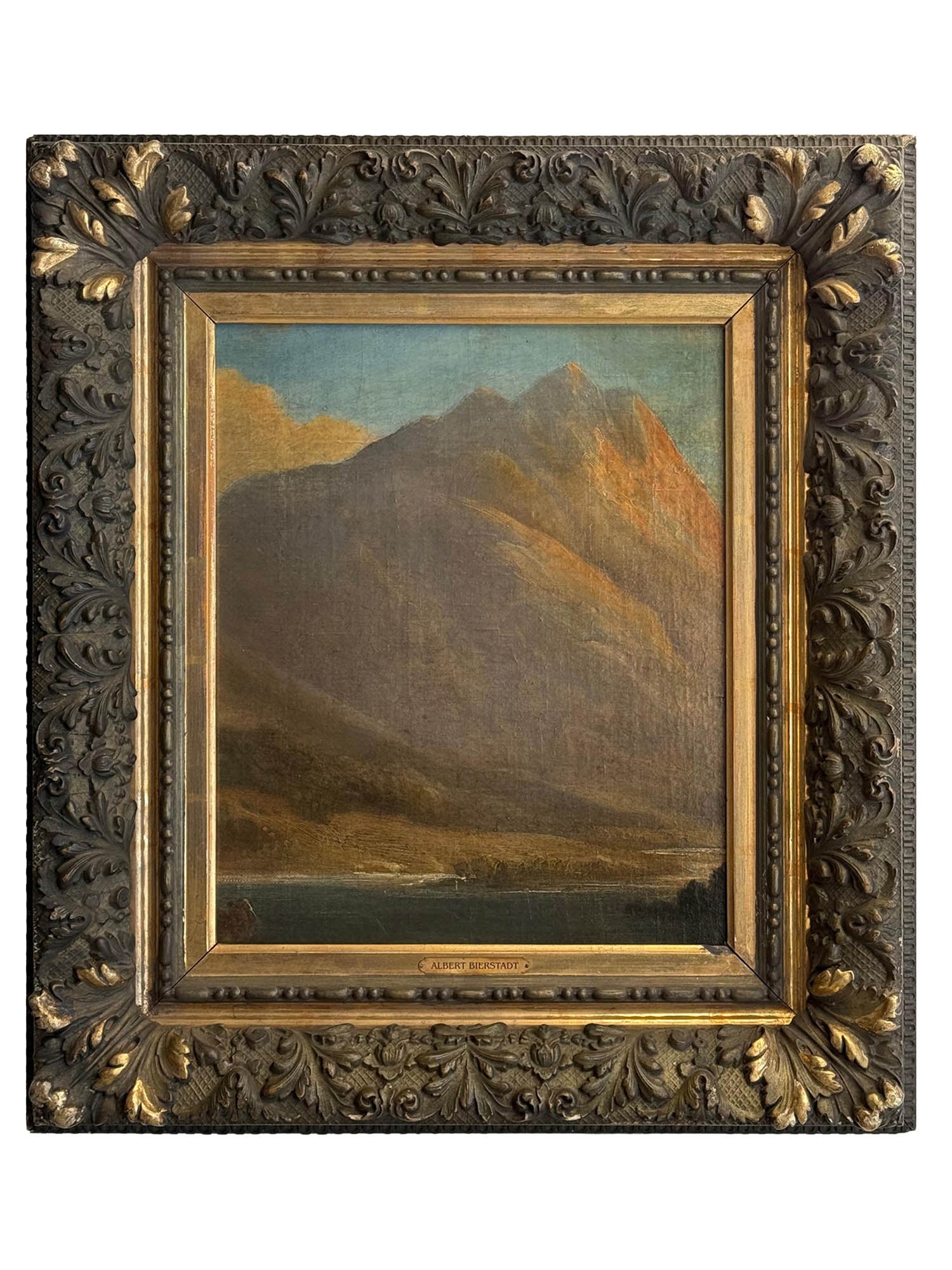 ATTRIBUTED TO ALBERT BIERSTADT (AMERICAN, 1830–1902) MOUNTAIN LANDSCAPE WITH LAKE: Oil on board. The composition depicts a monumental alpine mountain rising steeply above a tranquil lake in the foreground, the craggy peak illuminated by warm sunlight against a pale blue sky with sof