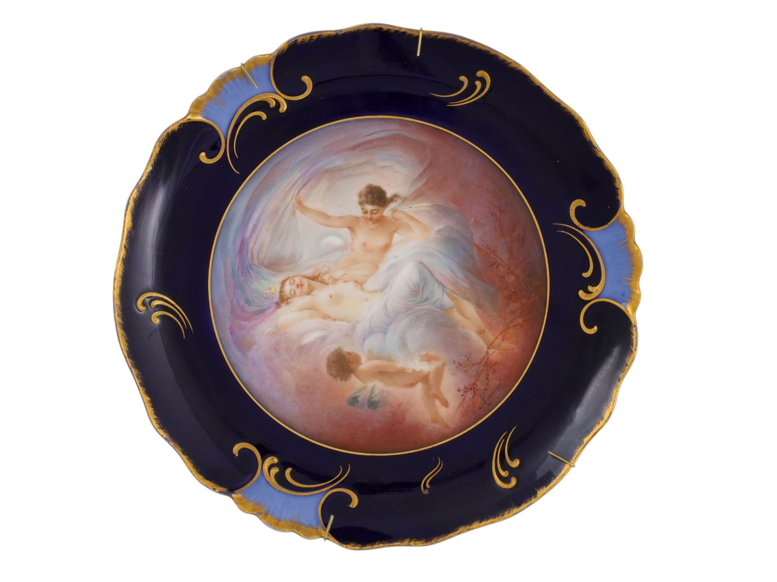 A LIMOGES HAND-PAINTED PORCELAIN CABINET PLATE, SIGNED NOUSTRE, LATE 19TH CENTURY: Of scalloped circular form with a cobalt blue ground, richly embellished with tooled gilt scrollwork along the rim, framing a finely painted central reserve. The scene depicts reclining classical maid