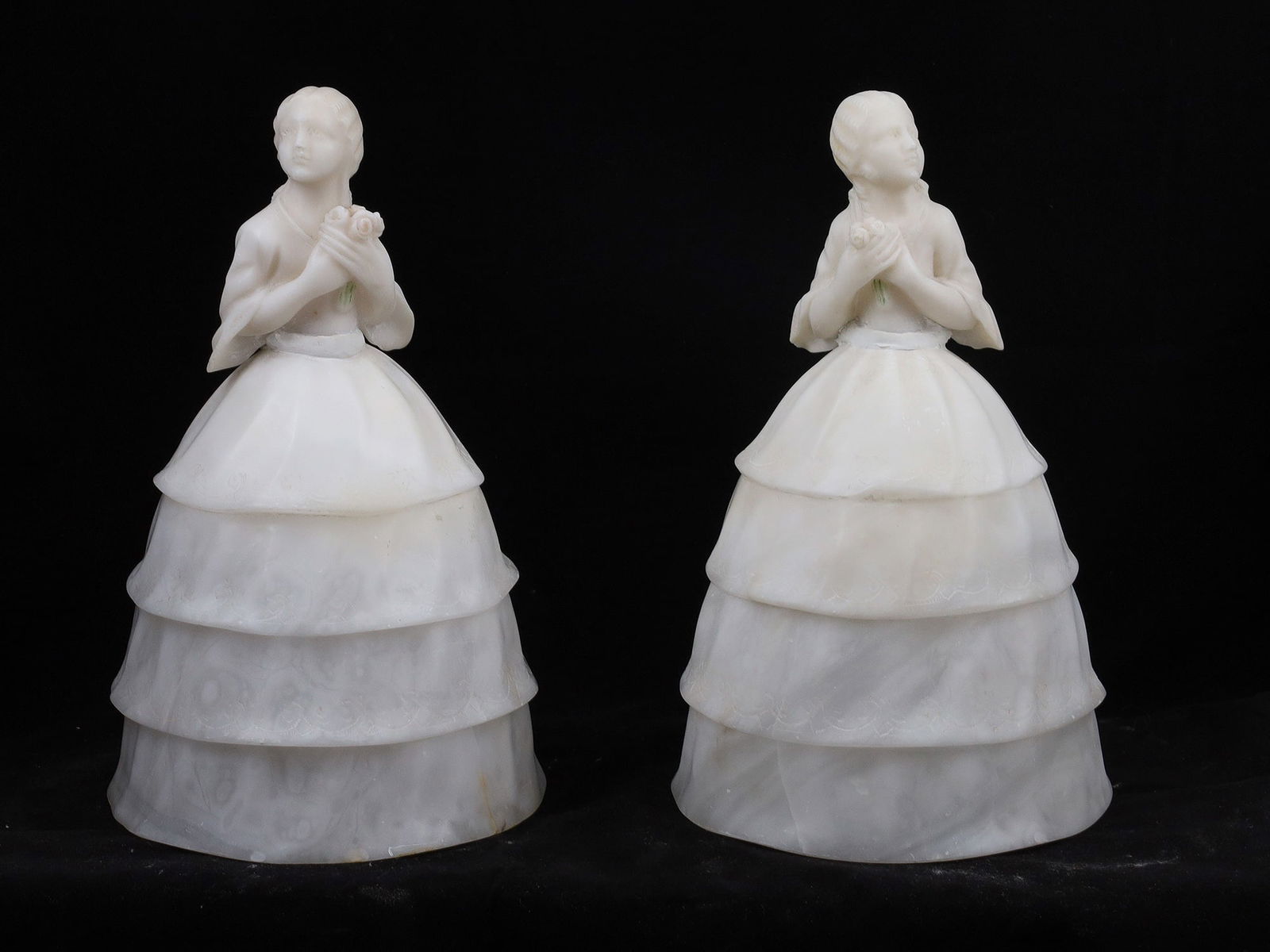 A PAIR OF ALABASTER FIGURAL TABLE LAMPS, 20TH CENTURY: Each modeled as a young woman in period dress, depicted holding a flower and with a tiered skirt forming the lampshade. Carved in translucent alabaster with subtle veining, the figures stand on circul