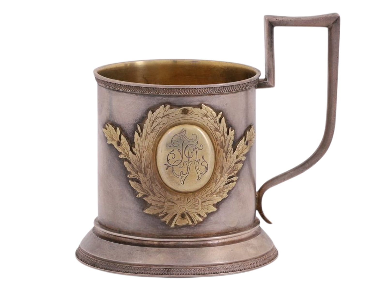 A RUSSIAN SILVER TEAGLASS HOLDER WITH INSCRIPTION, 19TH - 20TH CC.: A cylindrical tea glass holder with a rectangular handle, featuring a gilded cartouche in the shape of a laurel wreath with a bow. Inside the cartouche is an engraved monogram with initials. The upper