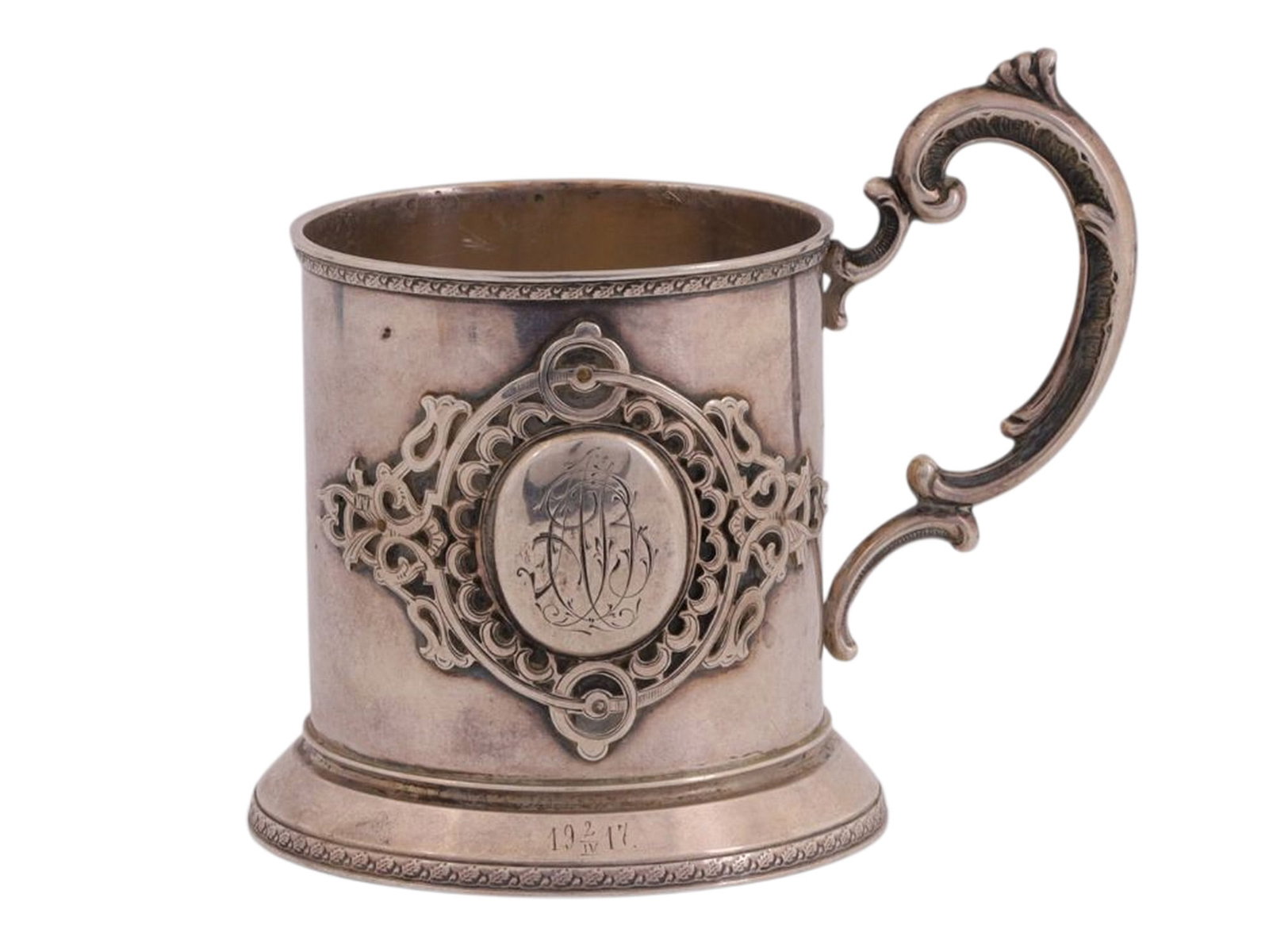 A RUSSIAN SILVER PODSTAKANNIK IN THE NEO-RUSSIAN STYLE, UNKNOWN WORKSHOP, LATE 19TH – EARLY 20TH: A cylindrical podstakannik (tea glass holder) with a rectangular handle, decorated with an applied cast silver ornamental cartouche in the Neo-Russian style. Within the cartouche is an engraved monogr