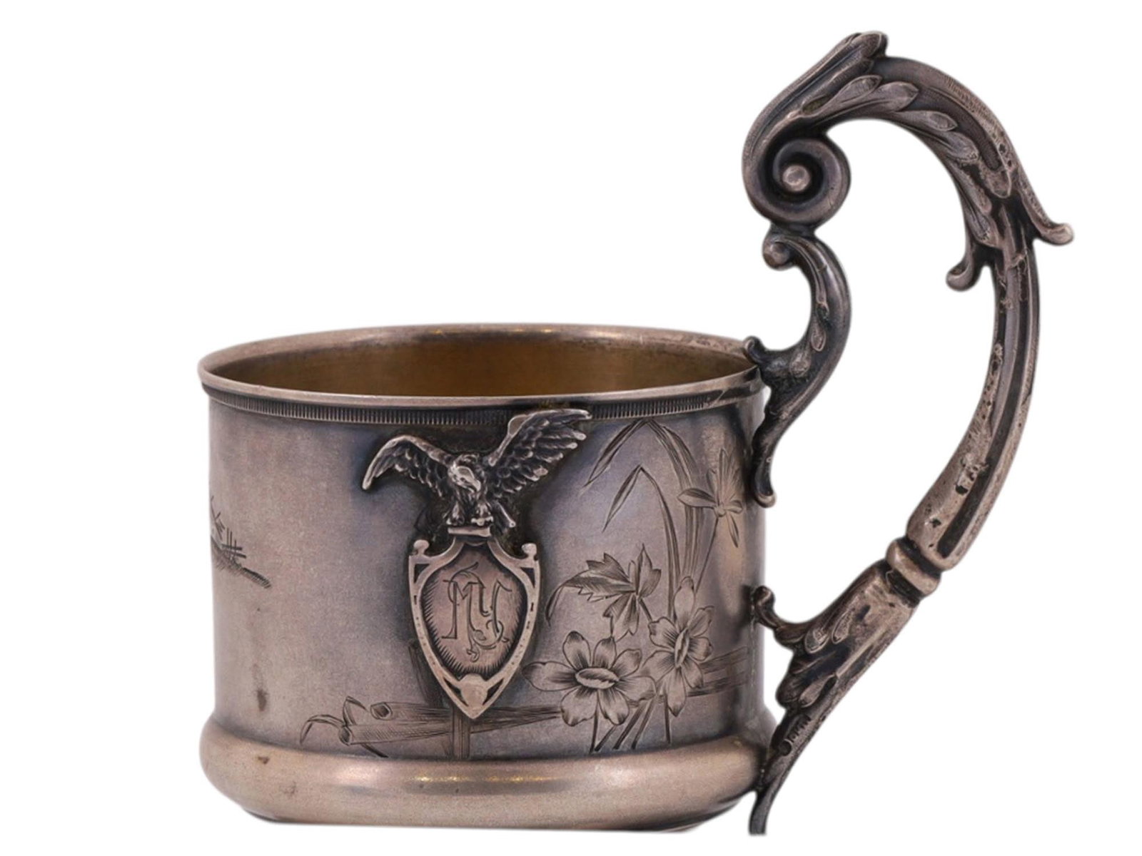 A RUSSIAN SILVER GIFT CUP WITH A RURAL LANDSCAPE, VASILY PULYATKIN WORKSHOP, MOSCOW, 1890S: A cylindrical tea glass holder tapering toward the base, featuring a rocaille handle and decorated on the side with an applied medallion depicting an eagle holding a shield. The shield is engraved wit