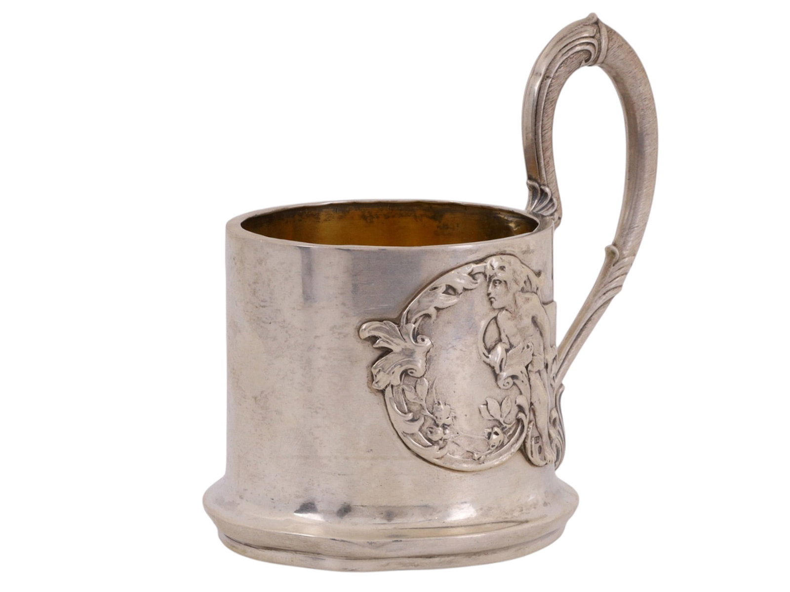 A RUSSIAN PARCEL-GILT SILVER TEA GLASS HOLDER WITH RELIEF: A Russian silver podstakannik (tea glass holder) in a transitional art-nouveau style, with a low relief image of a male youth among leaves and a curved handle. The inside with a smooth parcel-gilt fin