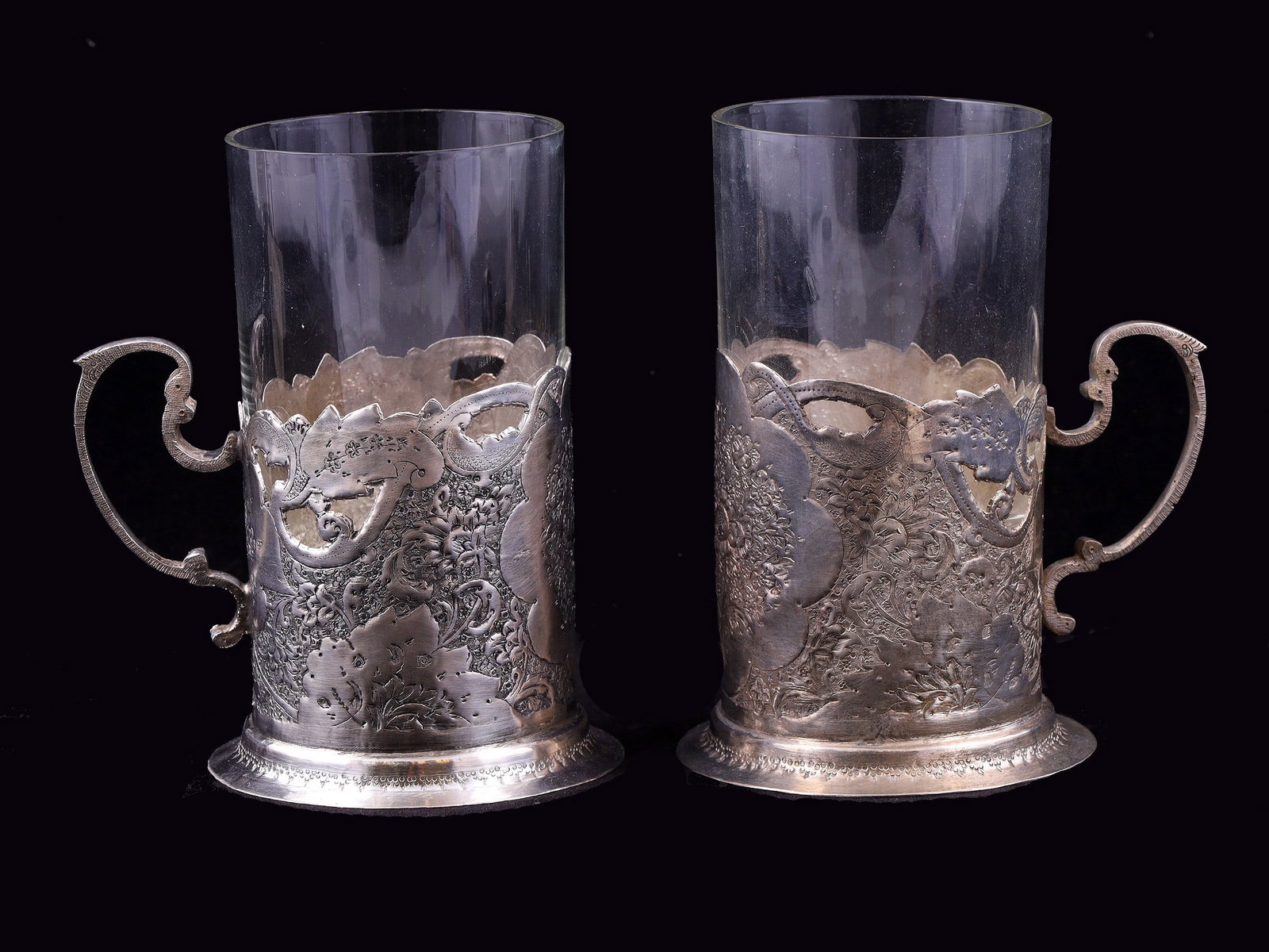A PAIR OF SILVER PODSTAKANNIKI, UNKNOWN WORKSHOP, RUSSIA, LATE 19TH CENTURY: Cylindrical podstakanniki with rocaille-form handles, shaped upper rims, and symmetrically engraved floral ornament highlighted with niello.The lower rims are decorated with a geometric guilloché