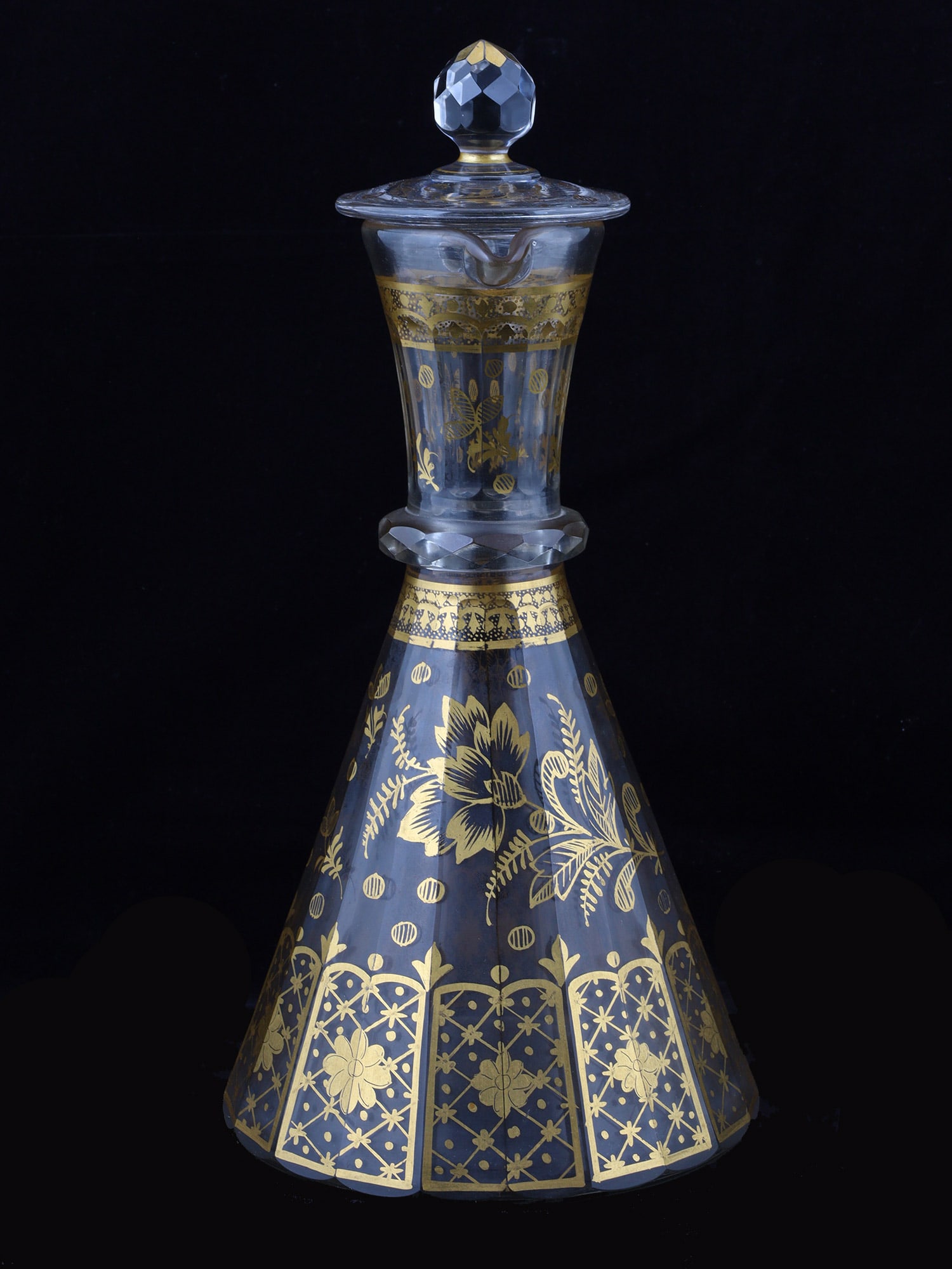 A RUSSIAN GILT-DECORATED CRYSTAL GLASS DECANTER BY THE NIKOLSKO-BAKHMETEV FACTORY, CA. 1860: Of tapering conical form, the faceted crystal body is decorated in gilt with stylized flowering branches, scattered circular motifs and a broad lower border of floral-filled latticework panels. The el