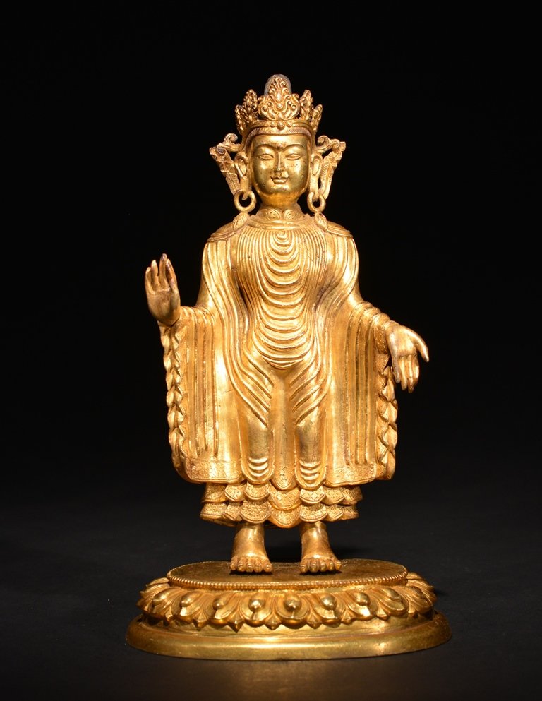 A GILT-BRONZE FIGURE OF BUDDHA: The standing figure is wearing a long pattern design traditional robe and hand in prayer position. The figure is wearing a tall royal crown with large earrings. There is a double vajra on the base of