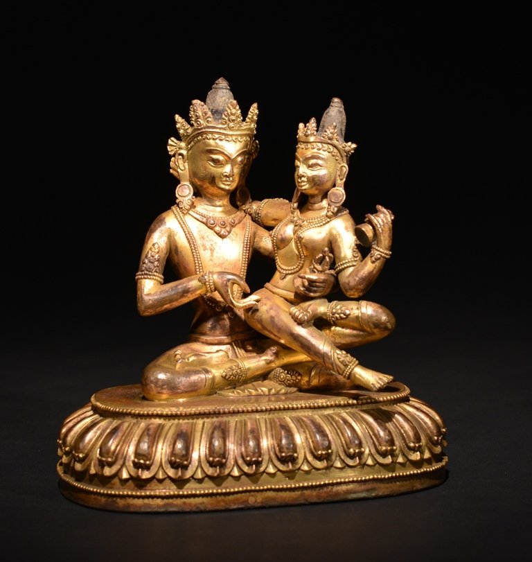 A GILT-BRONZE FIGURE OF VAJRADHARA WITH CONSORT: Seated in meditation on a double lotus pedestal with beaded borders, his left hand holding a vajra, his consort holds a vajra bell. 6 1/4 in. (15.9 cm) tall. Ming Dynasty