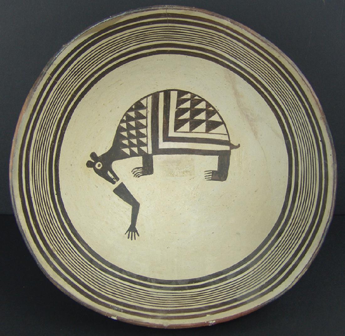 Mimbres Pottery Bowl: Fine, black-on-white bowl with multiple wide and fine line rim bands surrounding a bear (?) figure (who's mouth is near a human arm) it's half round body with geometric design and it's claws exposed. 