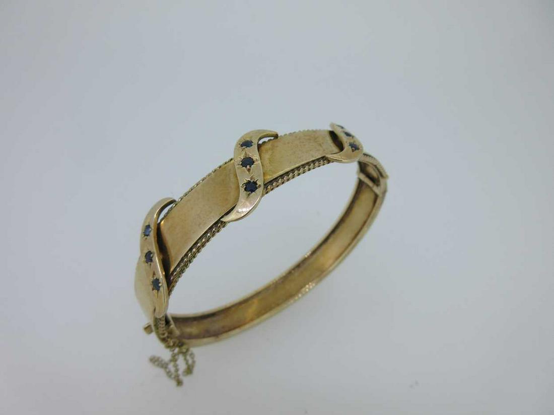 A hinged gold bangle set with sapphires,: the broad polished band with ropetwist edges on the front, crossed by three S figures each gypsy set with three round cut sapphires, hidden snap clasp, integral safety chain, London hallmarks for 9ct