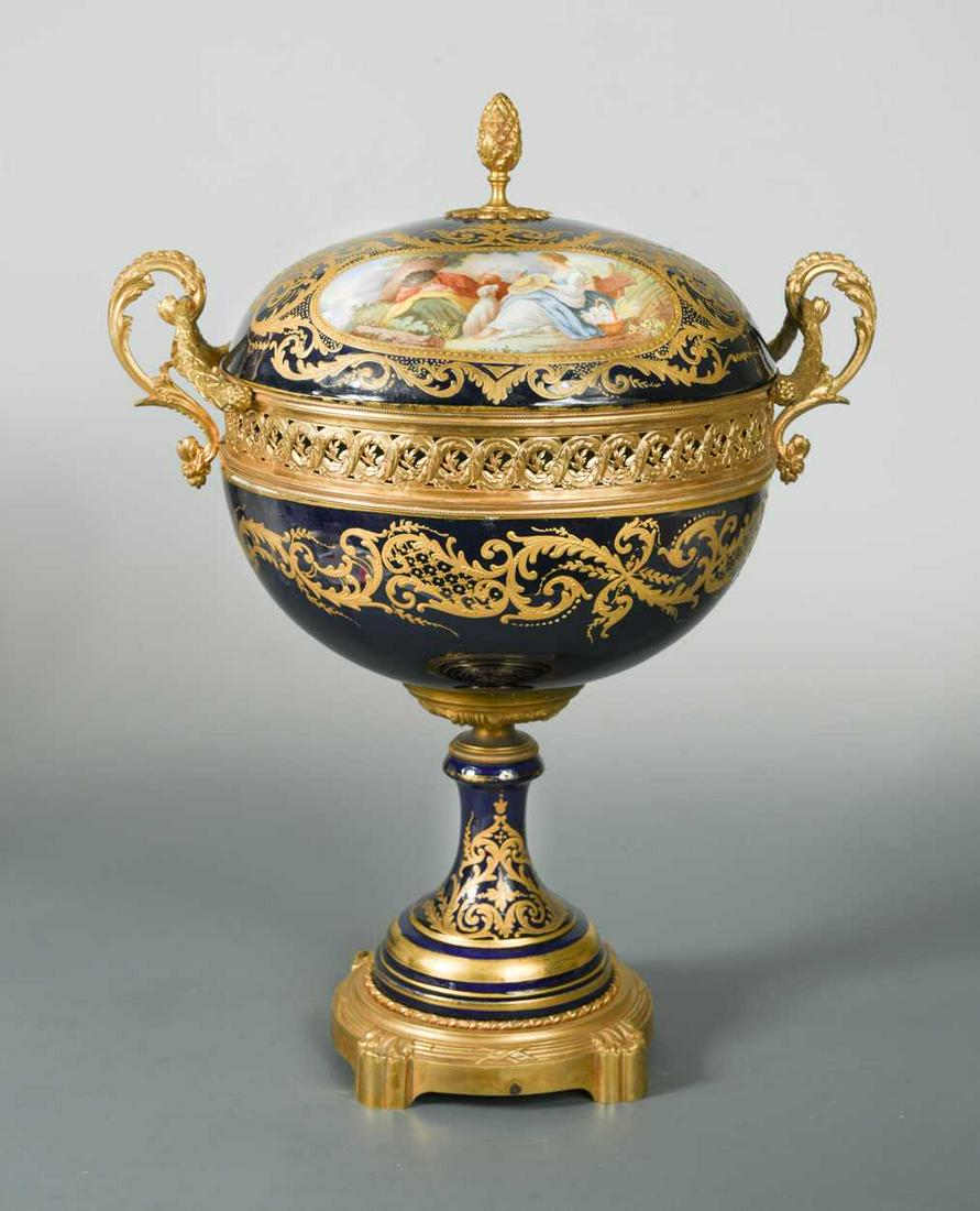 A Sevres two-handled ormolu mounted pedestal bowl and: the cover painted with two panels, one depicting a couple resting with a dog by a tree, the other with a house by a river, reserved on a blue ground body richly gilt with scrolling acanthus, the inter